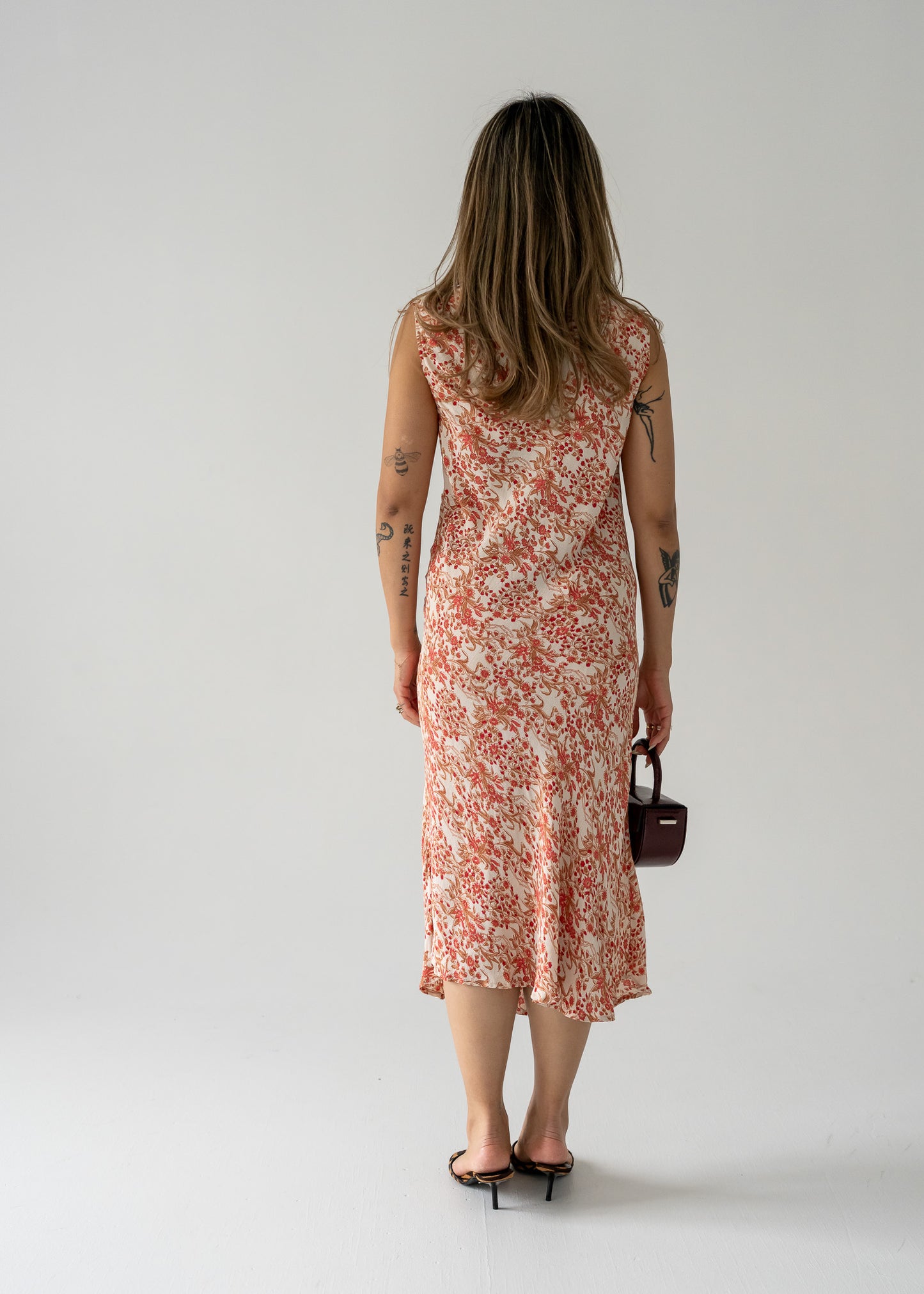Woman in a vintage sleeveless, cream and pink floral print midi dress holding a small bag against a plain background