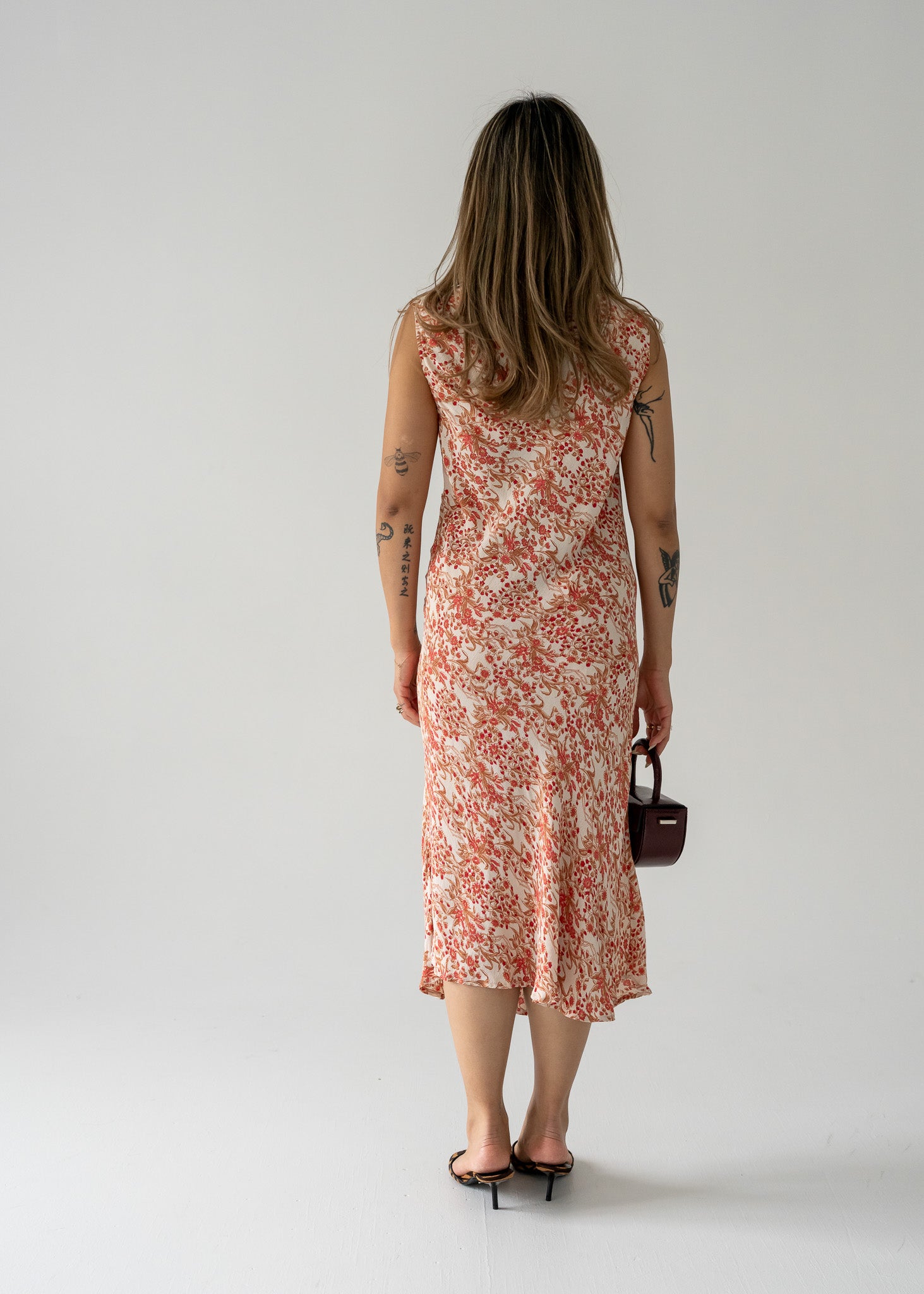 Woman in a vintage sleeveless, cream and pink floral print midi dress holding a small bag against a plain background