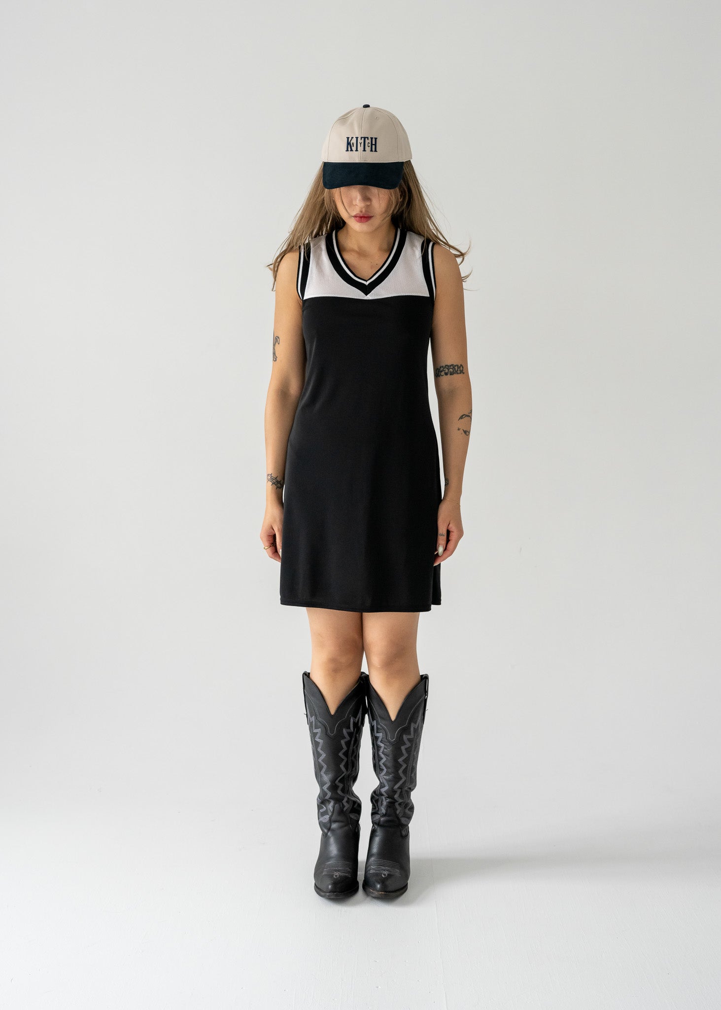 A model wearing a black and white mesh vintage 90's sporty tank dress with a rounded v neck, sleeveless design, and wearing cowboy boots and a hat.