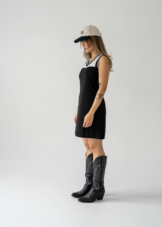 Woman in black and white mesh vintage 90's sporty tank dress with a rounded v neck and cowboy boots with a cap, standing against a plain background.