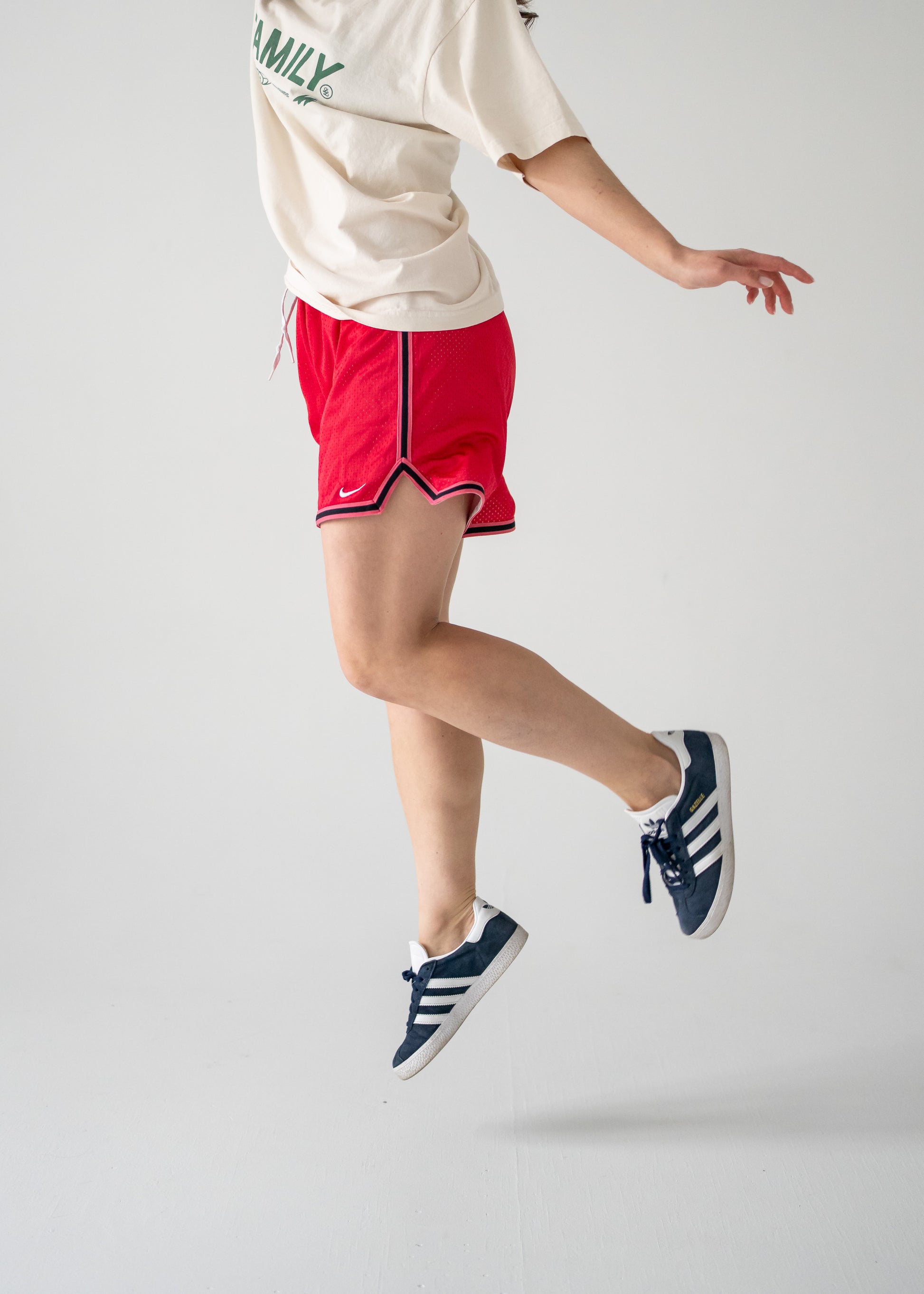 A person is mid-jump wearing vintage red mesh Nike shorts with black side stripes and a white top, paired with blue sneakers.