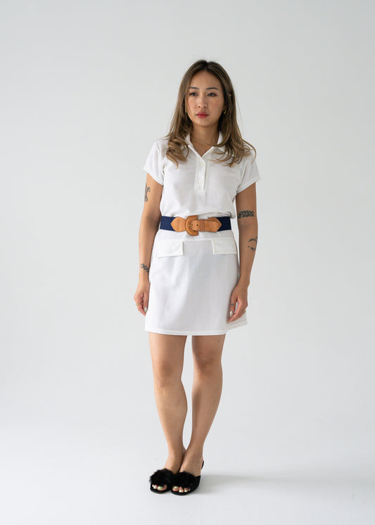 A woman standing against a white background wearing a vintage bright white drop waist button up collared short sleeve tennis mini dress with a brown belt and black shoes.
