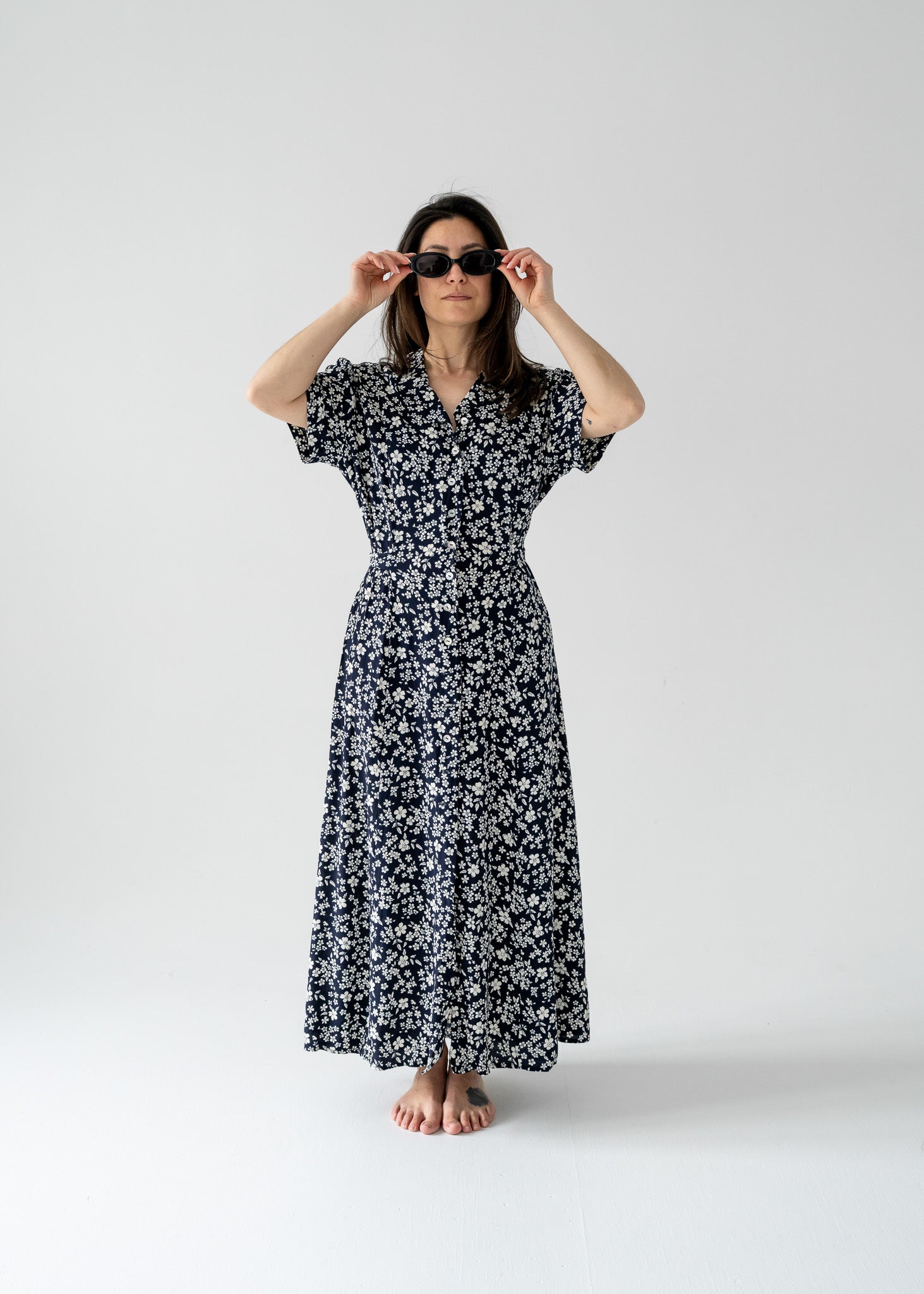A model wearing a vintage navy and white cherry blossom print button-up short sleeve maxi dress with a V-neck, long rounded collar, and buttons ending 18 inches from the hem.