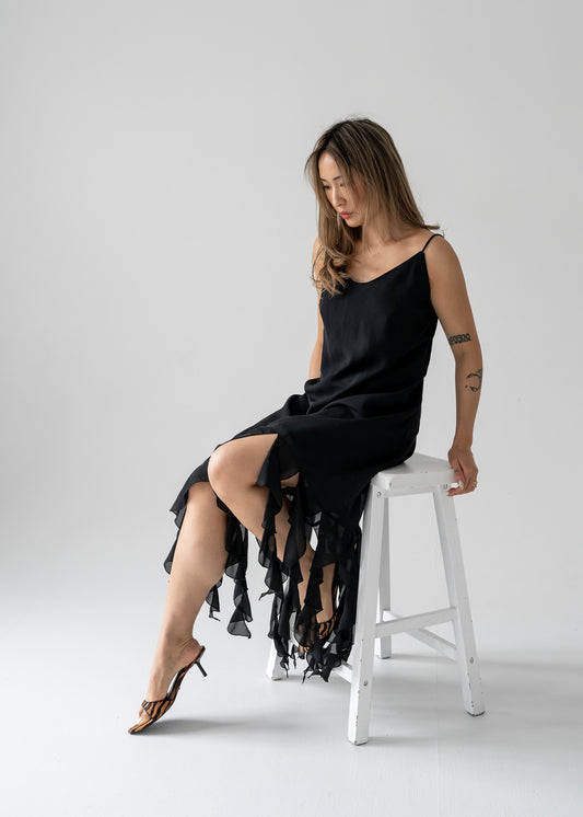 A woman seated on a white stool wearing a vintage black long dress with a trailing ruffle frill hem and thin straps.