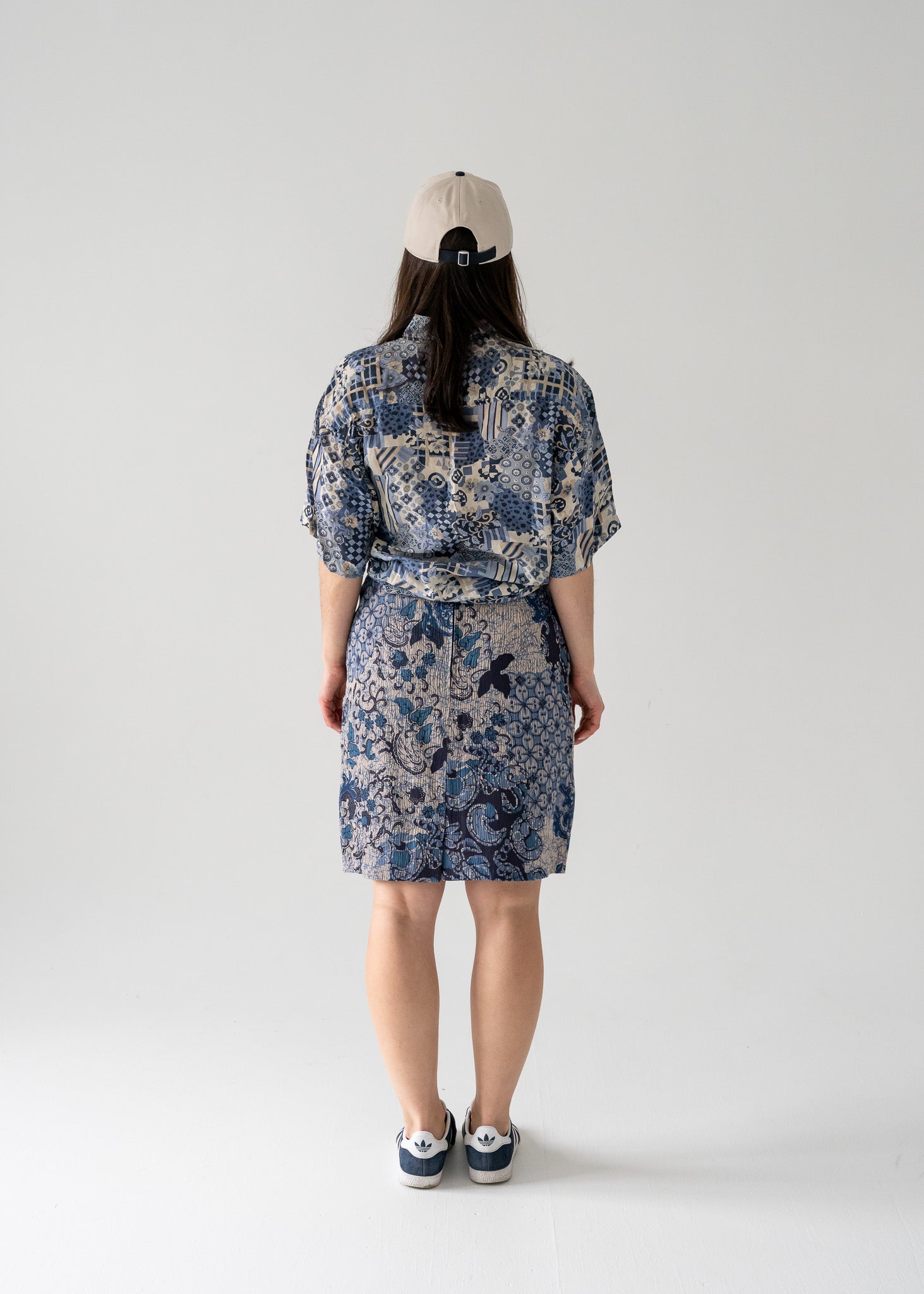 Person wearing a knee-length blue floral pattern vintage pencil skirt, patterned silk shirt and cap, standing against a plain background.