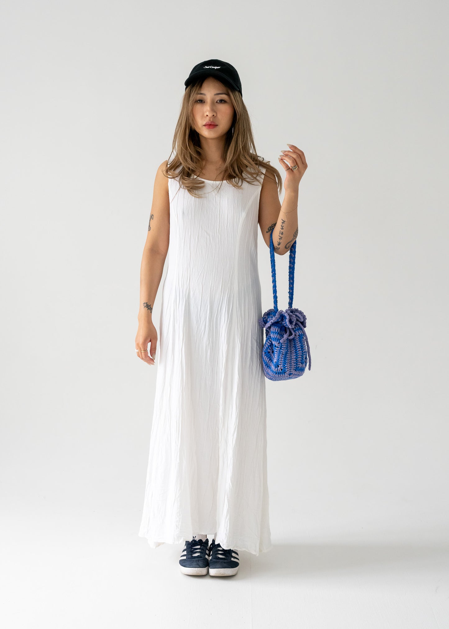 A woman standing against a white background wearing a vintage ivory crinkle lightweight floor-length princess seam sleeveless dress with a cap, holding a blue bag, and wearing flat shoes.