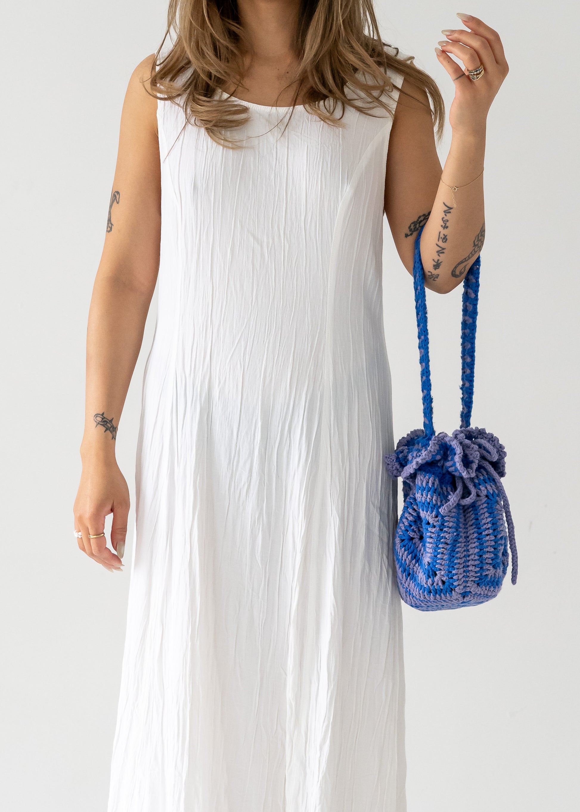 A person wearing a vintage sleeveless, ivory, crinkle texture floor-length dress with a princess seam, holding a blue woven bucket bag.