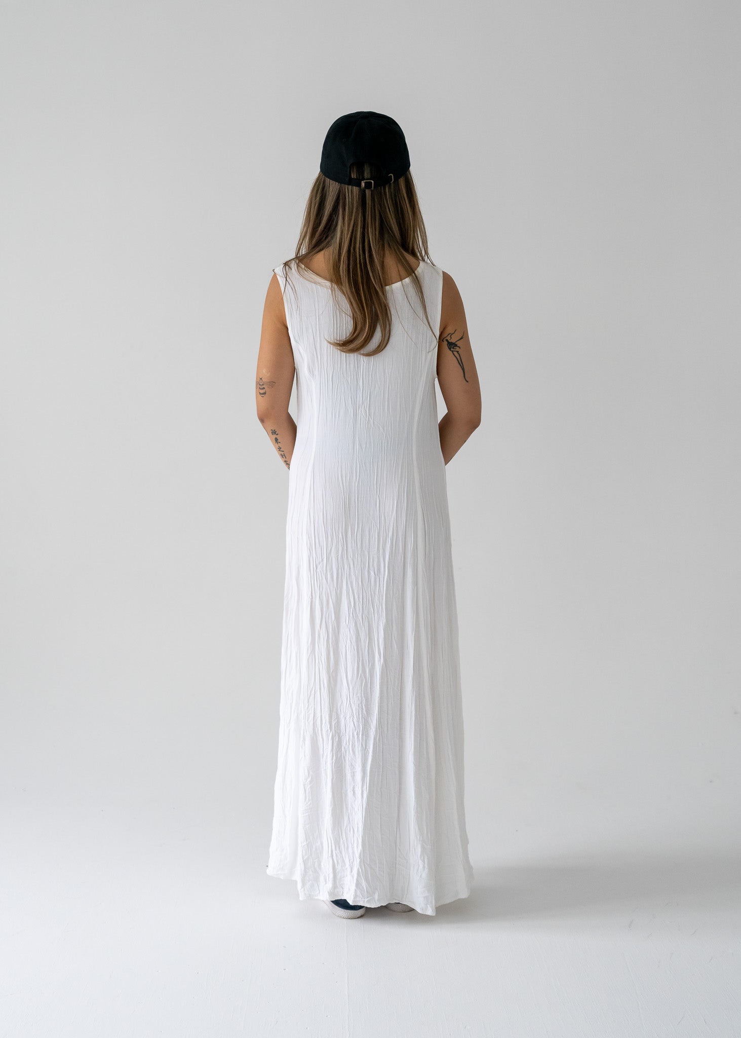 Person wearing a vintage ivory crinkle lightweight floor-length princess seam sleeveless dress and black cap on a plain background