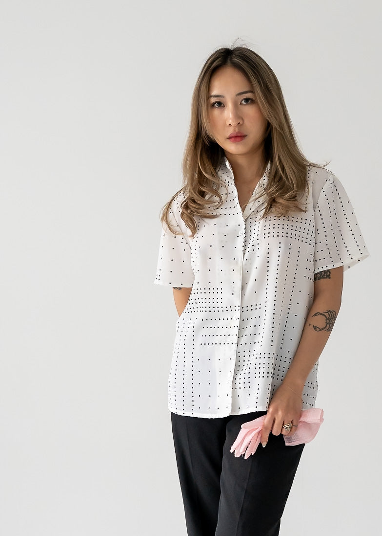 A woman wearing a vintage white short sleeve button up blouse with black square polka dots, paired with black pants.