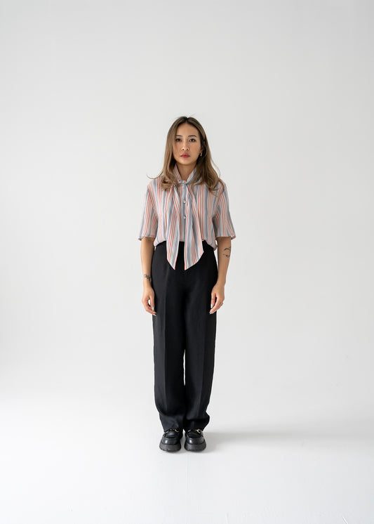 A woman standing against a white background wearing a multi-colored thin vertical striped sheer tie neck short sleeve button up blouse in shades of orange, purple, and white on a light blue base, paired with dark pants.