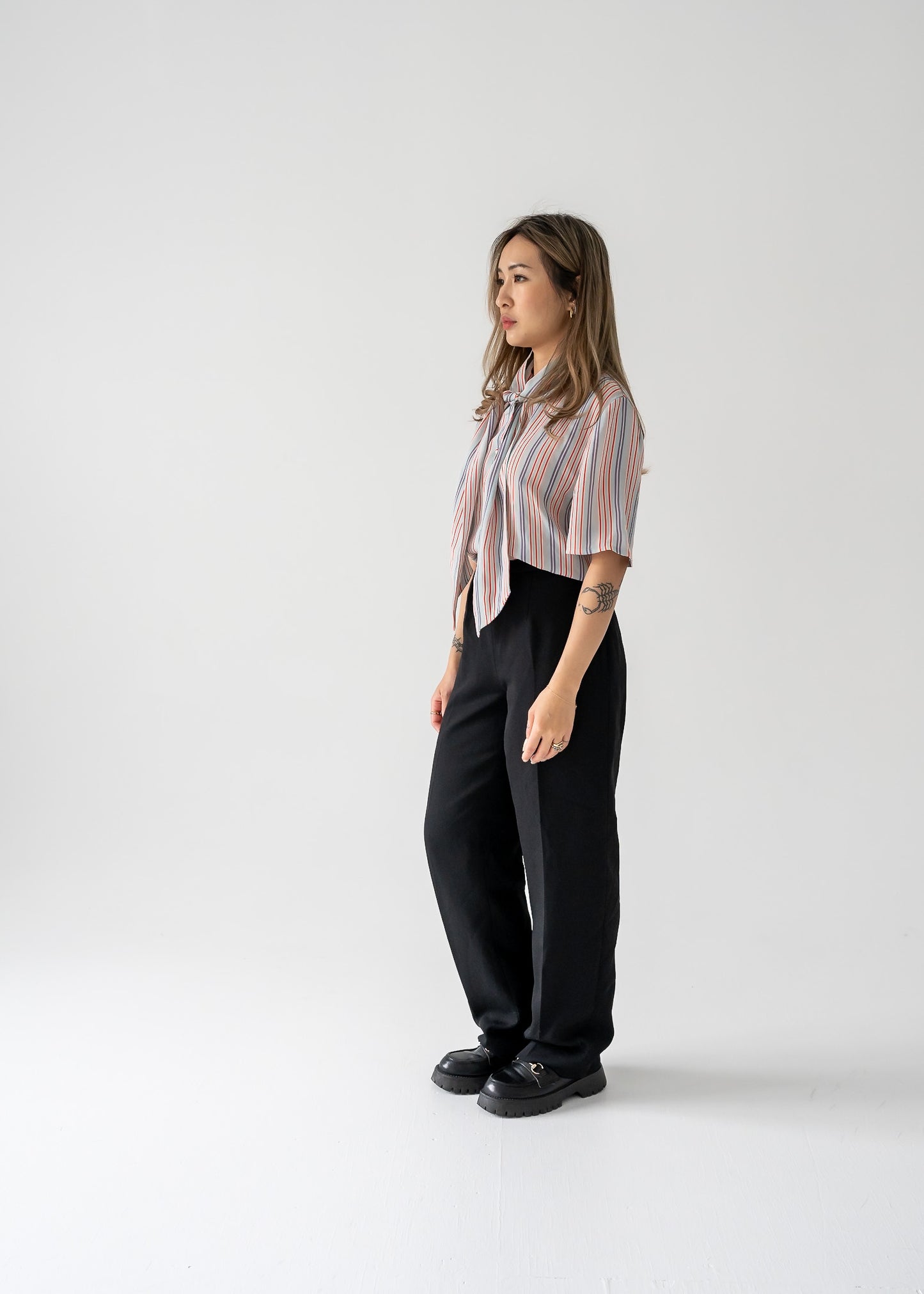 A woman standing against a white background wearing a multi-colored thin vertical striped sheer tie neck short sleeve button up blouse in shades of orange, purple, and white on a light blue base, paired with dark pants.