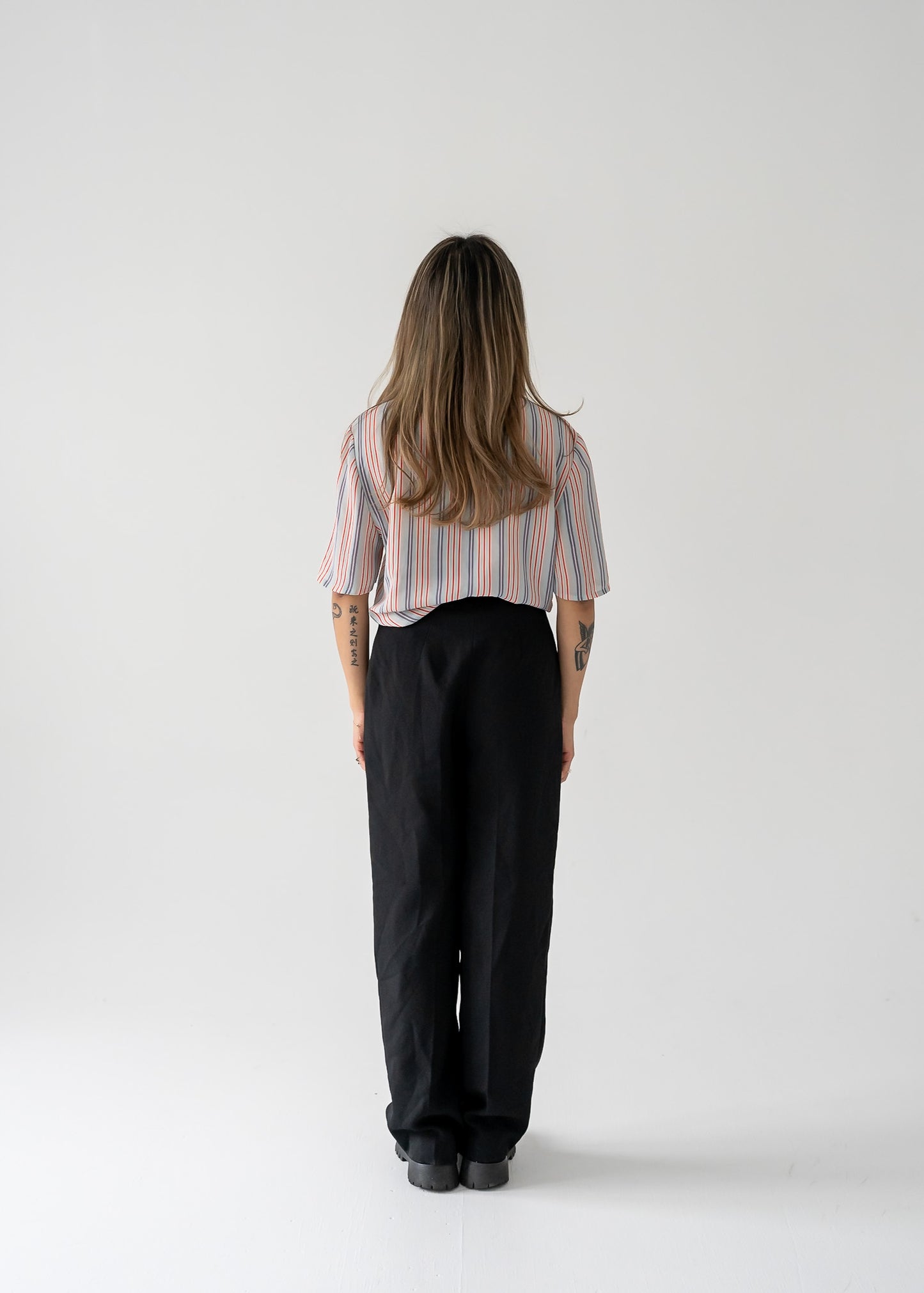 A woman standing against a white background wearing a vintage multi-colored thin vertical striped sheer tie neck short sleeve button up blouse in shades of orange, purple, and white on a light blue base, paired with dark pants.