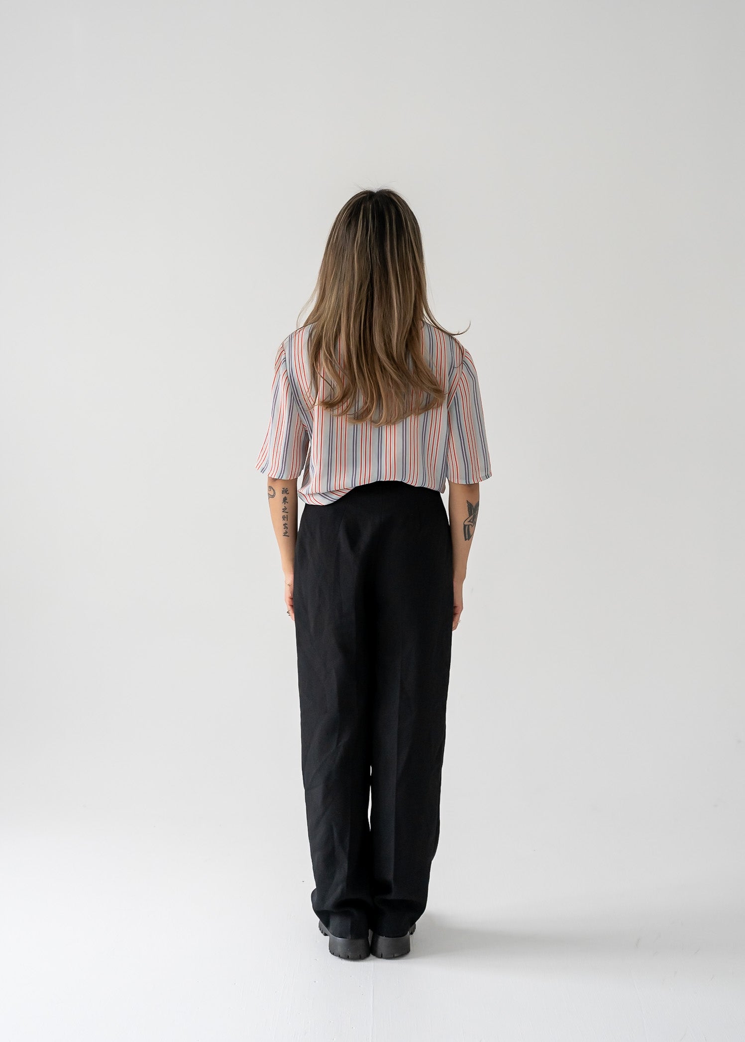 A woman standing against a white background wearing a vintage multi-colored thin vertical striped sheer tie neck short sleeve button up blouse in shades of orange, purple, and white on a light blue base, paired with dark pants.