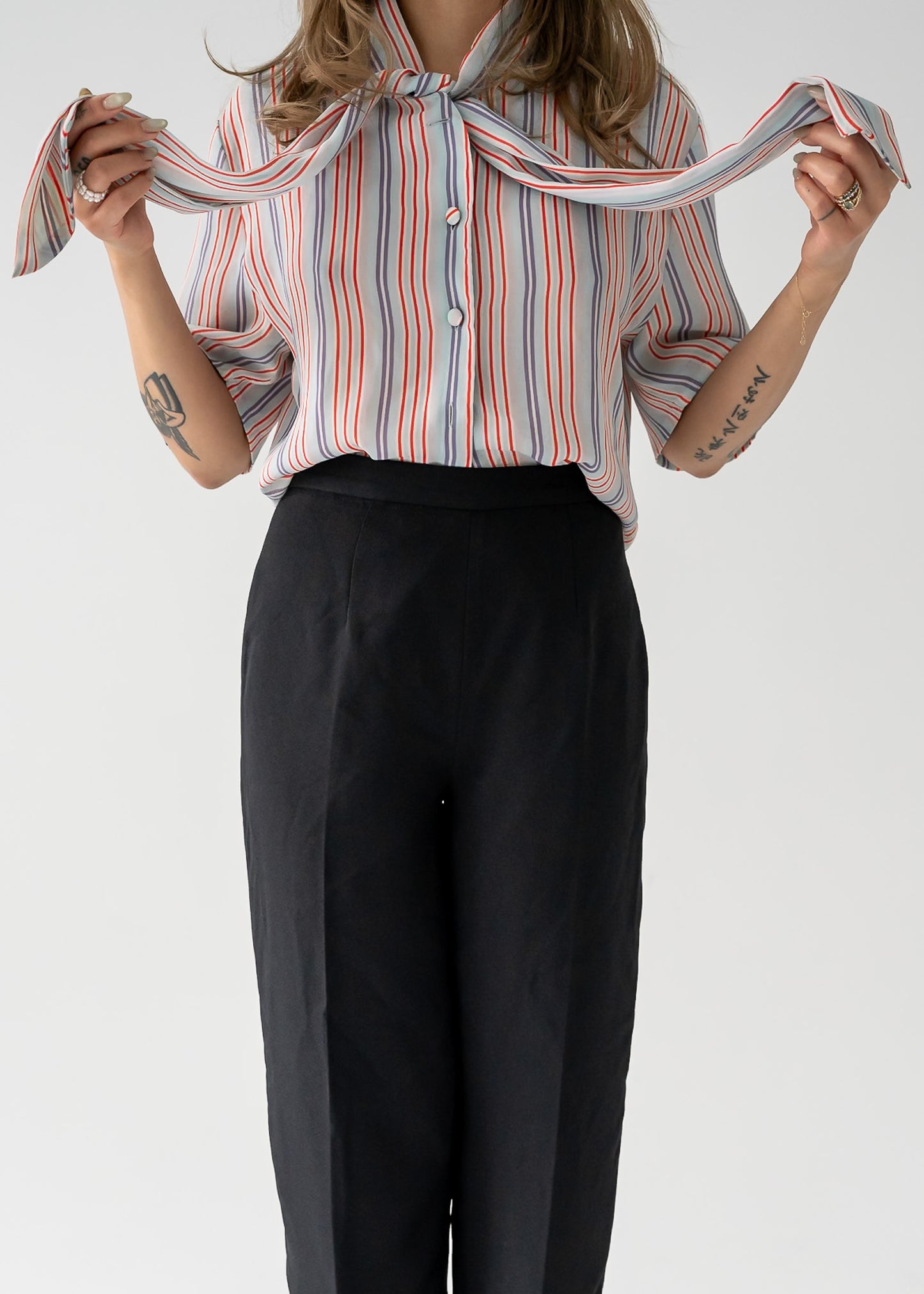 A woman standing against a white background wearing a multi-colored thin vertical striped sheer tie neck short sleeve button up blouse in shades of orange, purple, and white on a light blue base, paired with dark pants.