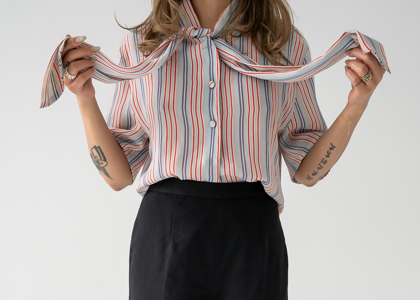 A woman standing against a white background wearing a multi-colored thin vertical striped sheer tie neck short sleeve button up blouse in shades of orange, purple, and white on a light blue base, paired with dark pants.