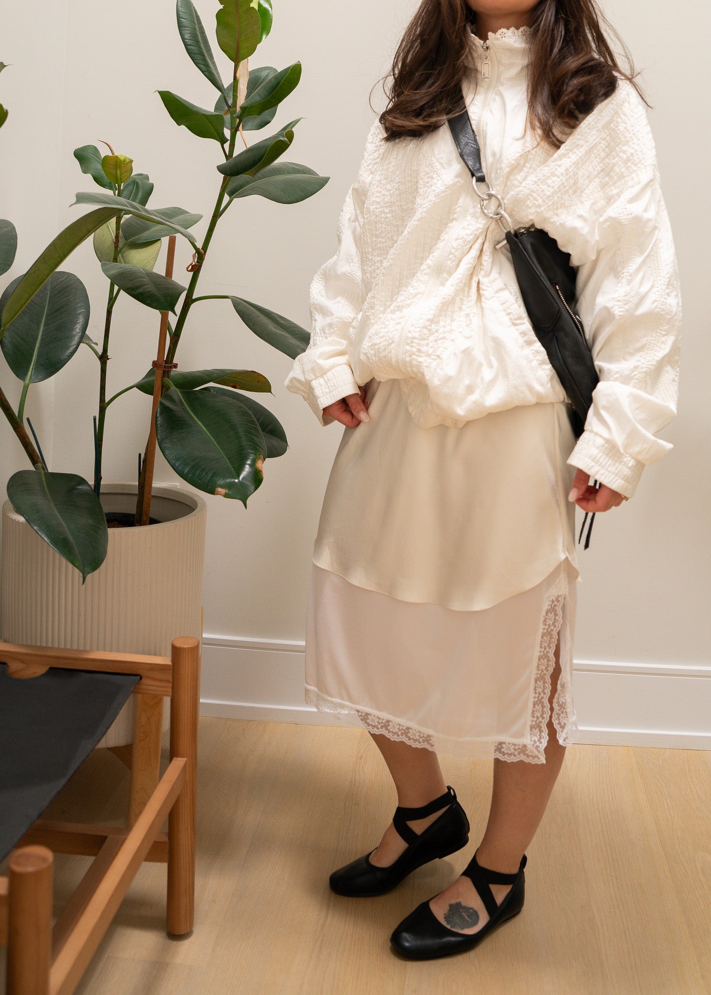 Person wearing a white outfit with a black bag in a room with a plant and wooden shelf.