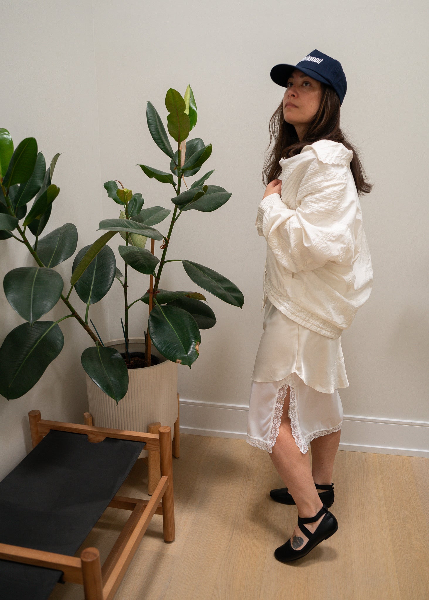 Person wearing a white jacket, slip and lace skirt and blue cap standing next to a plant indoors.