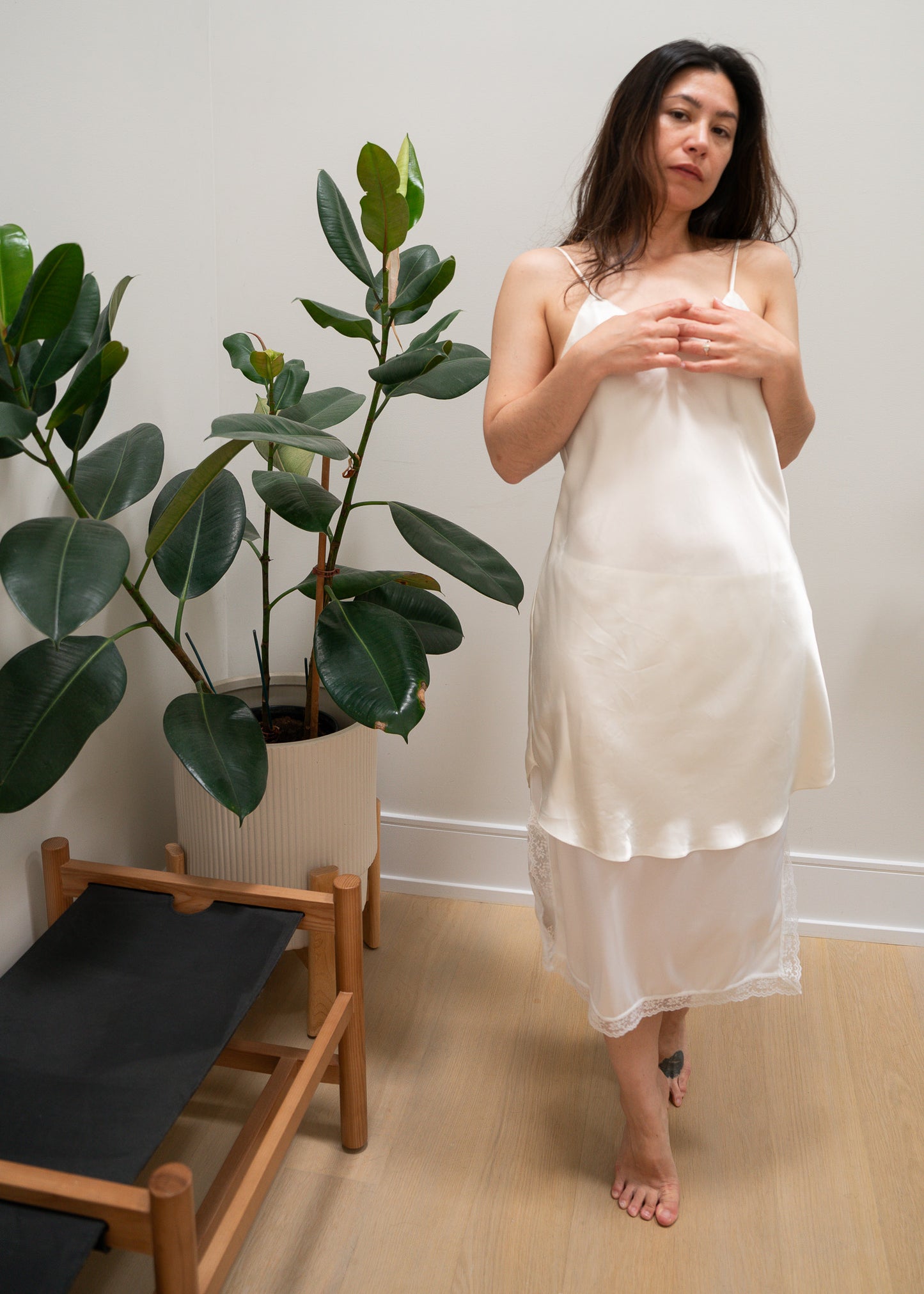 Woman in a white slip standing in a room with a plant and chair.