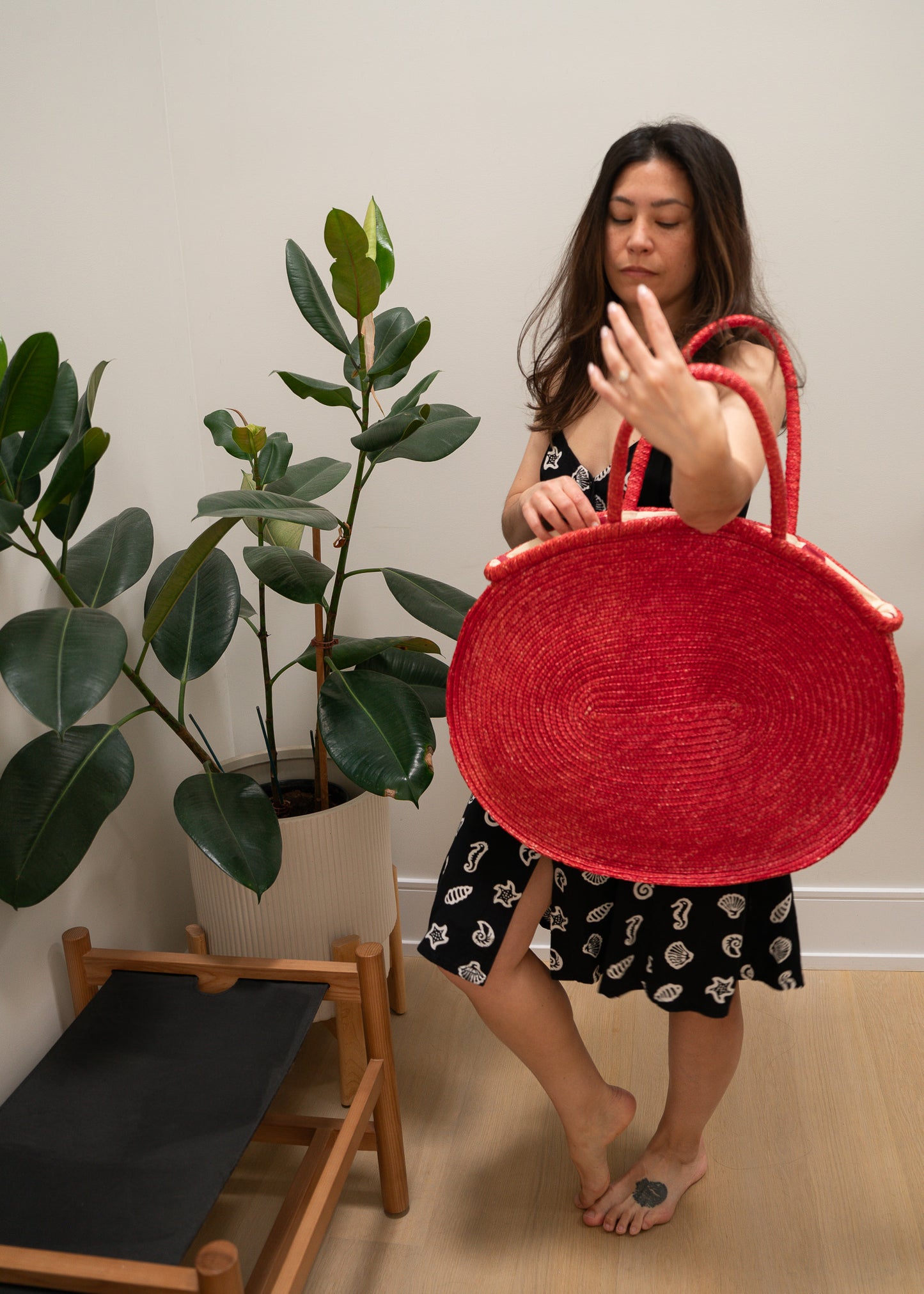Woman holding a red woven bag indoors with a plant and chair in the background