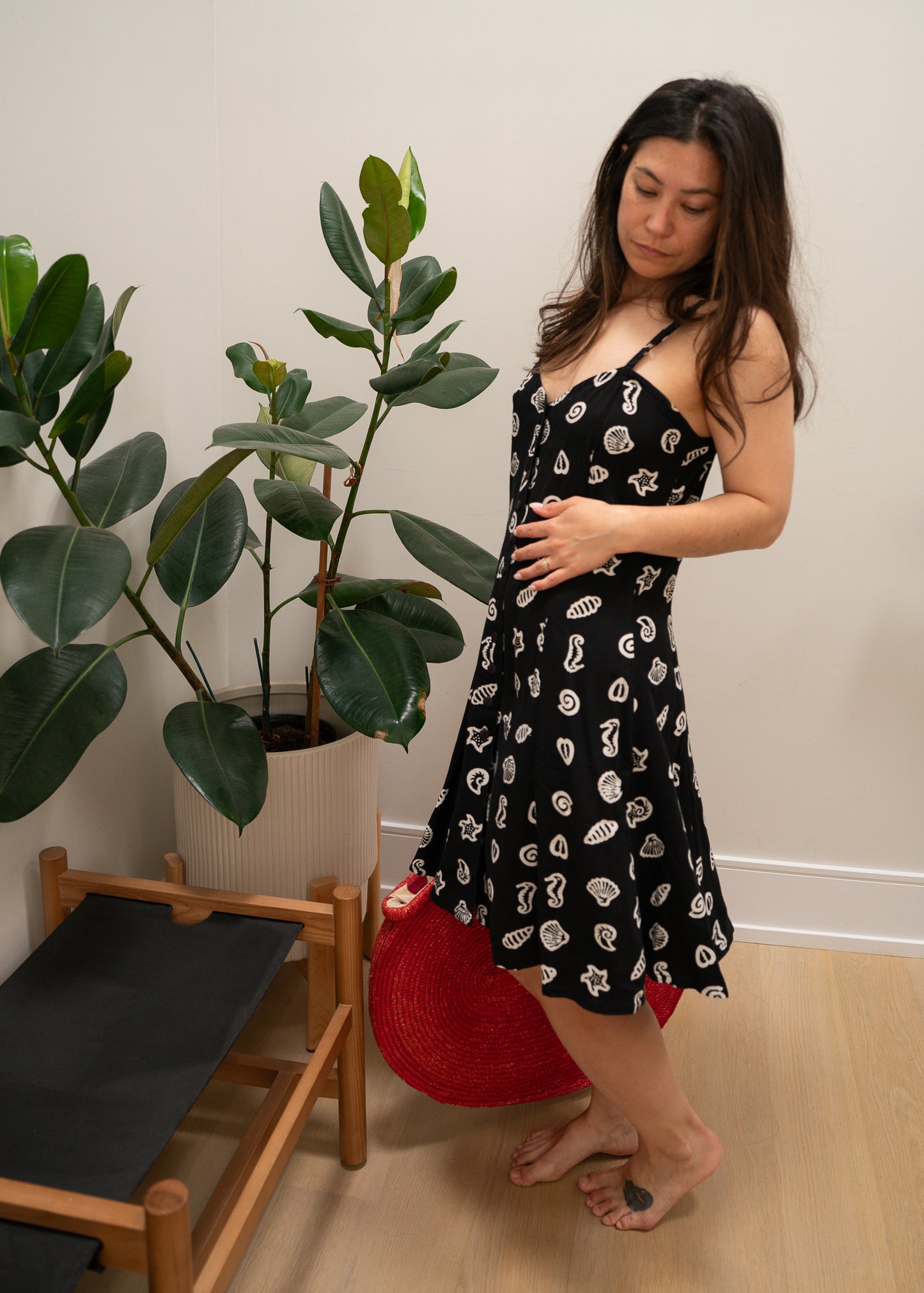 Woman in a black with white ocean pattern dress standing next to a plant indoors