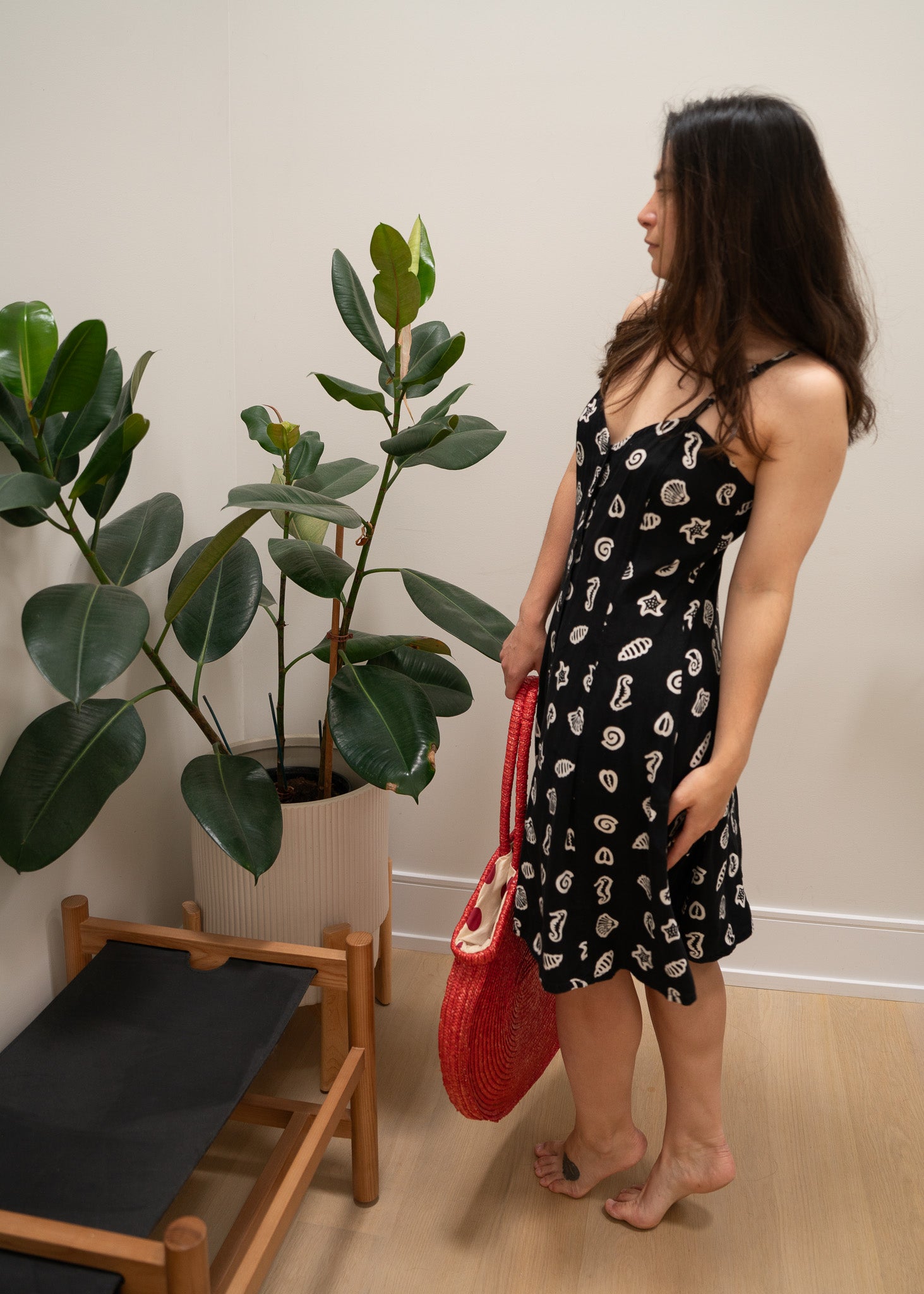Woman in a black and white ocean patterned dress holding a red bag indoors next to a plant.