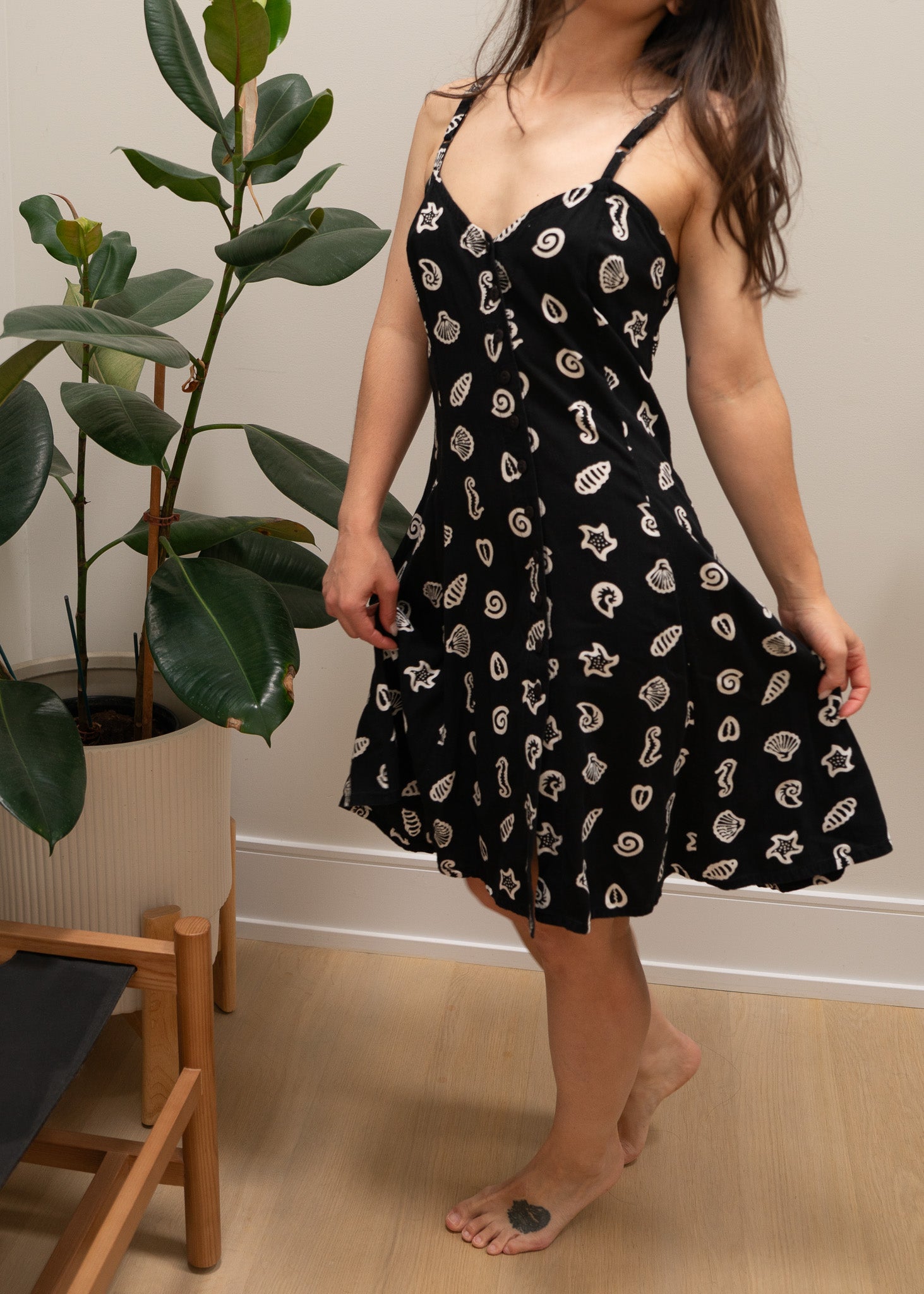 Person wearing a black dress with white ocean patterns in a room with a plant and wooden chair.
