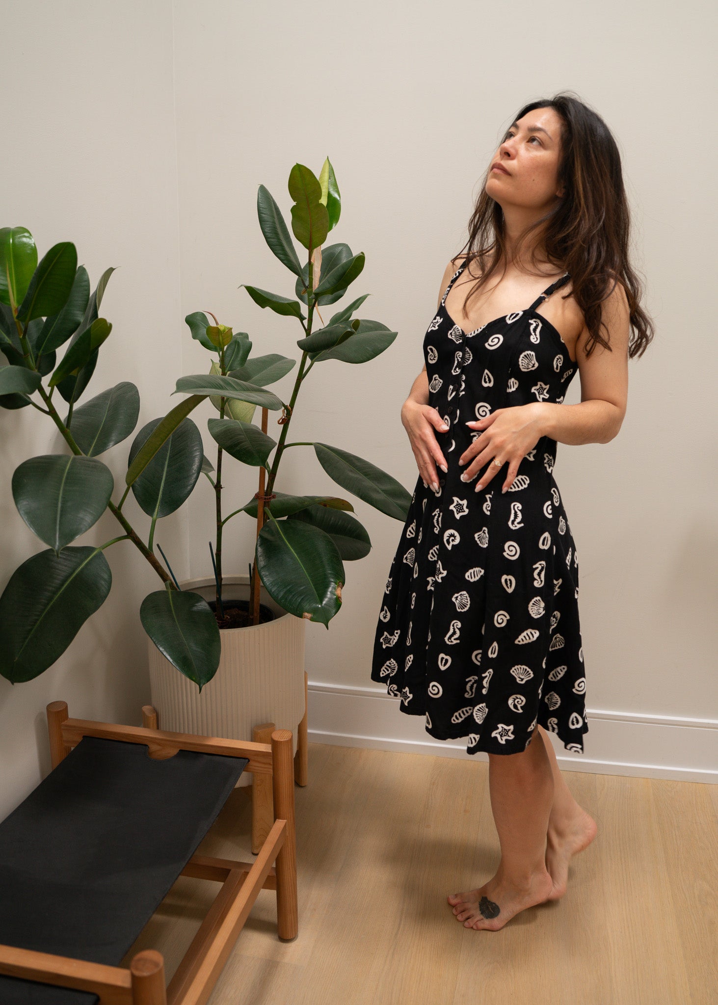 Woman in a black dress with white sea patterns standing indoors next to a plant.