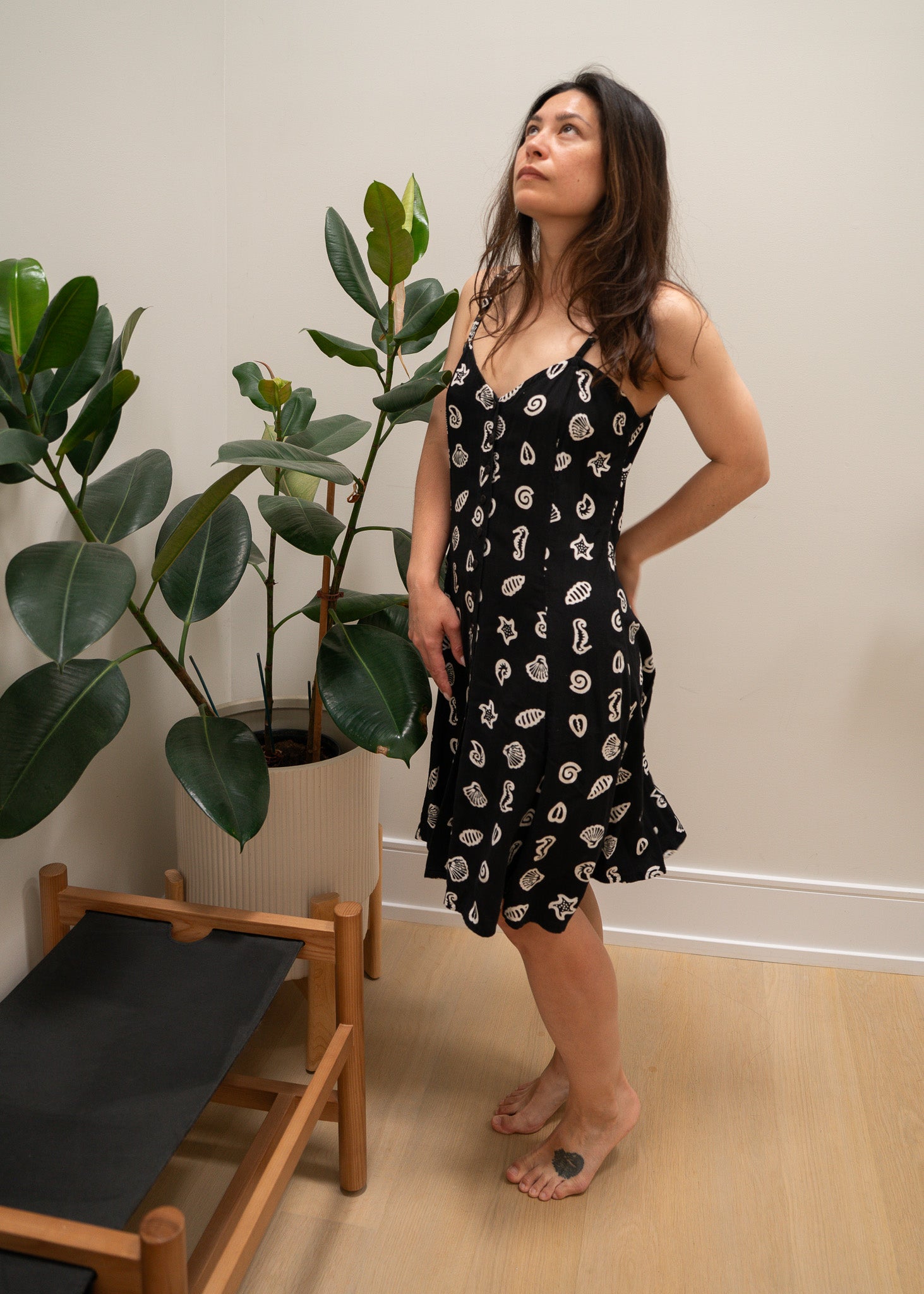 Woman in a black with white sea patterned dress standing next to a plant indoors.