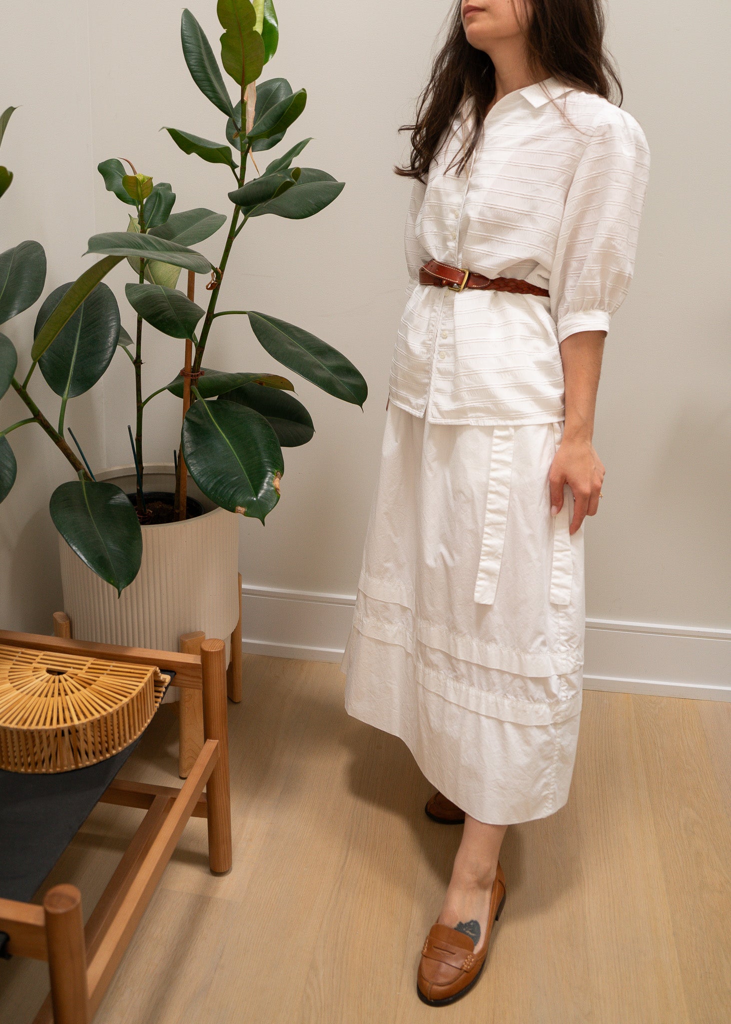 Woman in a white white striped puffy sleeve blouse, leather belt and white skirt standing in a room with a plant and wooden chair.