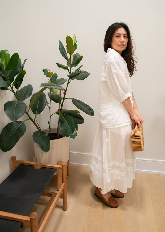 Woman in a white puffy sleeve blouse and white skirt standing in a room with plants and a chair.