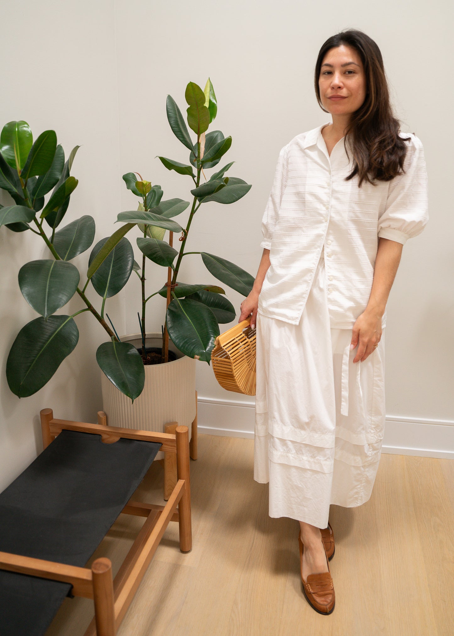 Woman in a white white striped puffy sleeve blouse and white skirt standing in a room with plants and a chair.