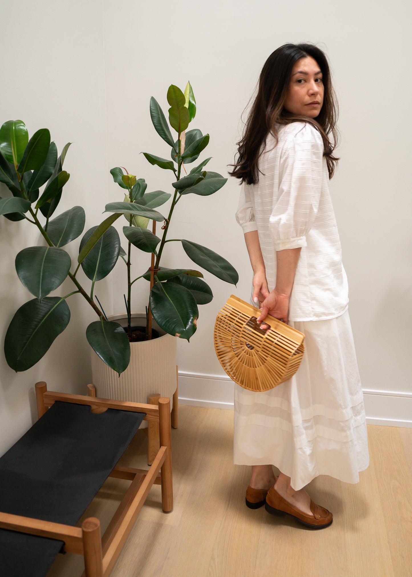 Woman in a white white striped puffy sleeve blouse and white skirt holding a wooden bag in a room with plants and a chair.