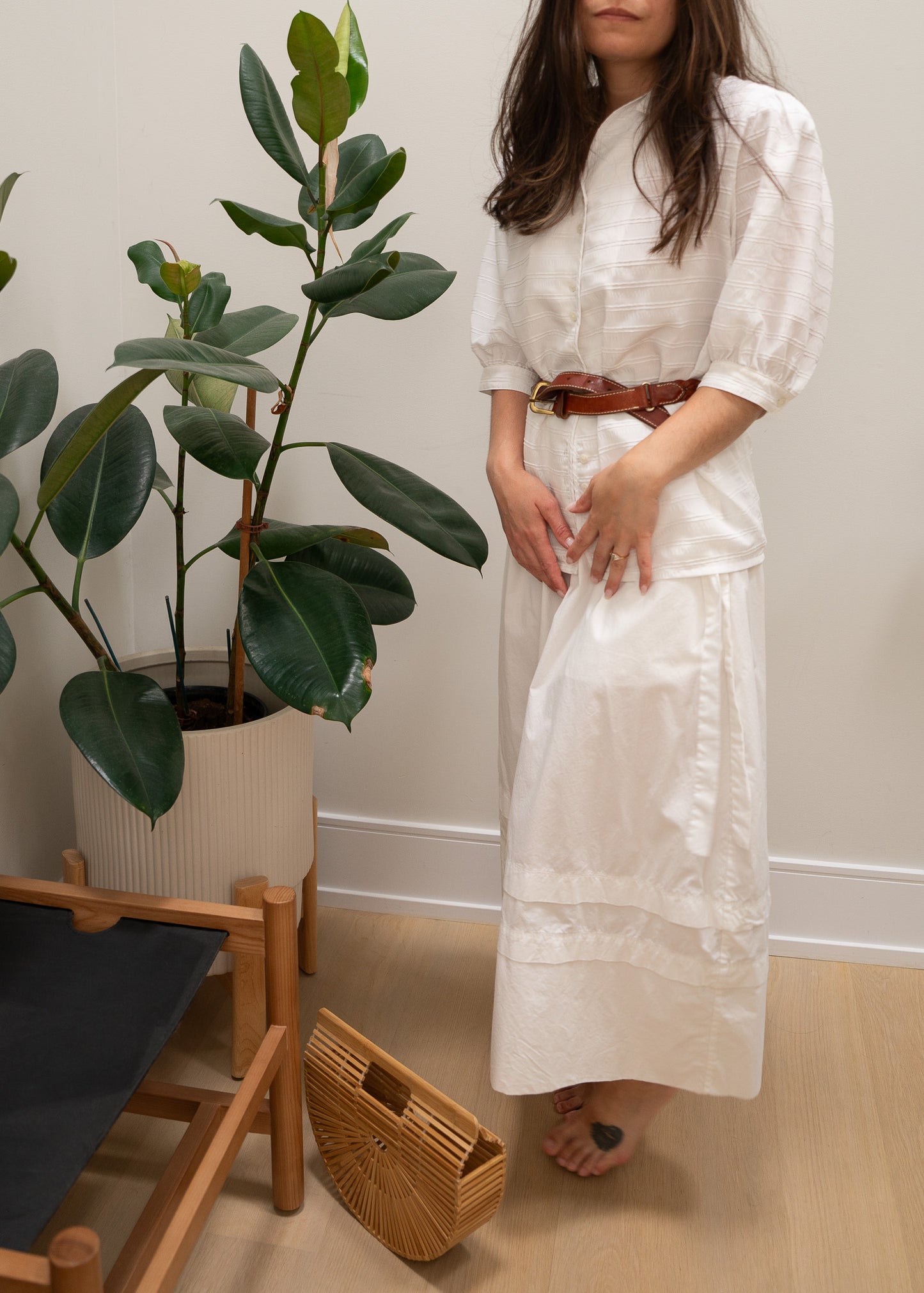 Woman in a white white striped puffy sleeve blouse, leather belt and white skirt standing next to a plant and chair indoors.