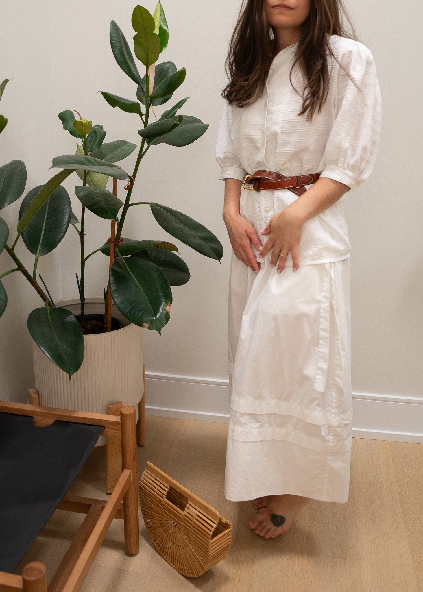 Woman in a white white striped puffy sleeve blouse, leather belt and white skirt standing next to a plant and chair indoors.