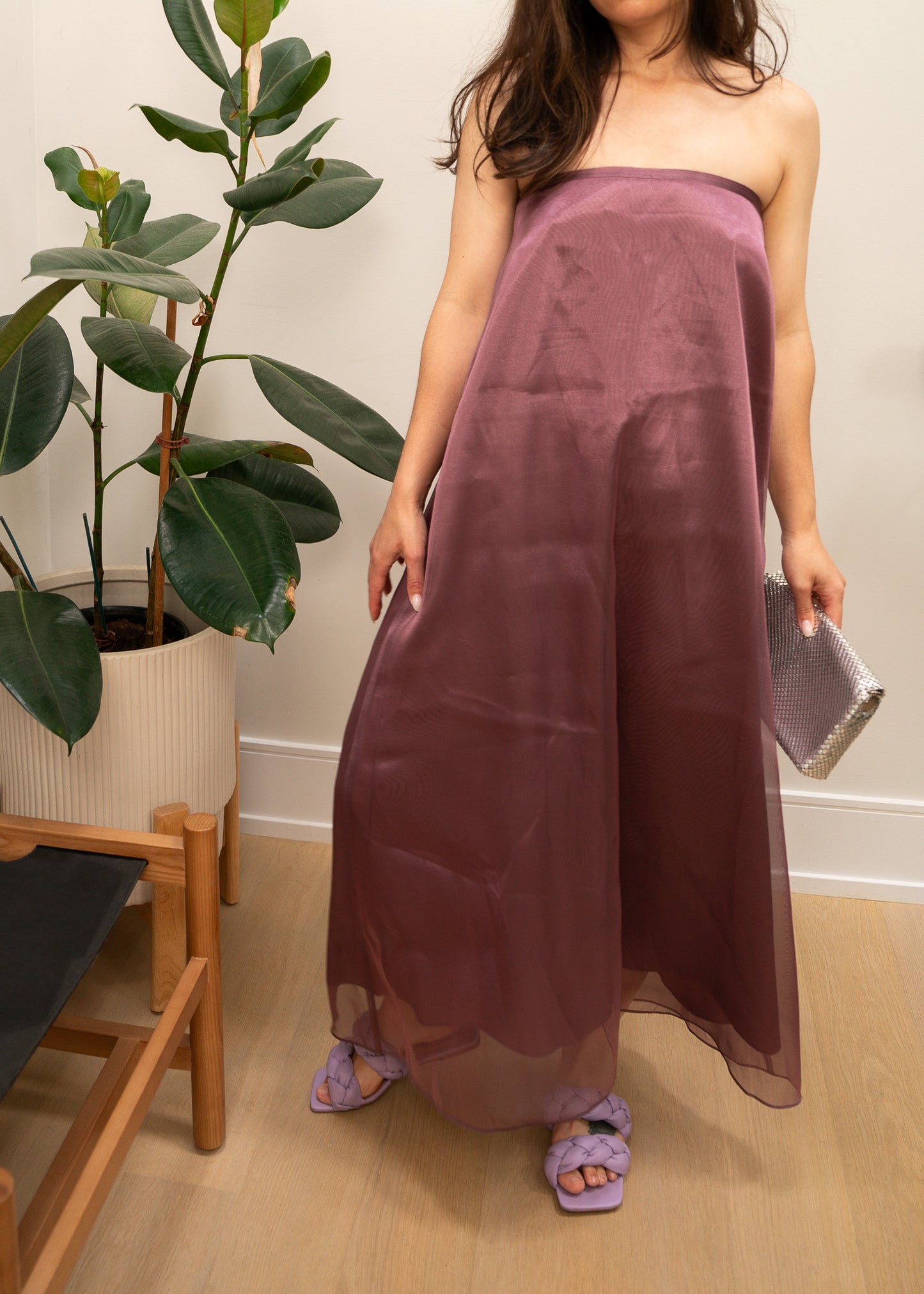 Woman wearing a strapless mauve purple dress holding a clutch in a room with a plant and wooden chair.
