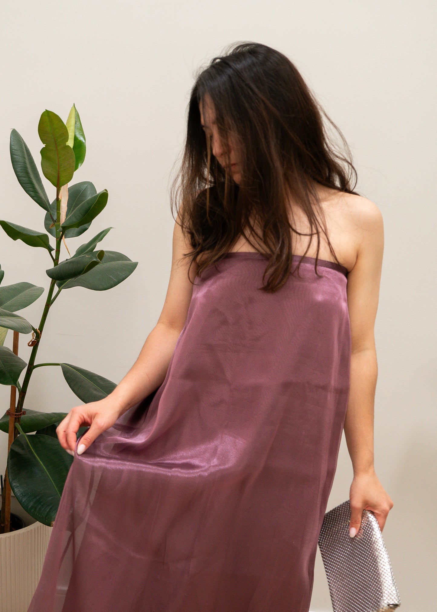 Woman wearing a mauve purple strapless dress standing next to a plant against a plain background