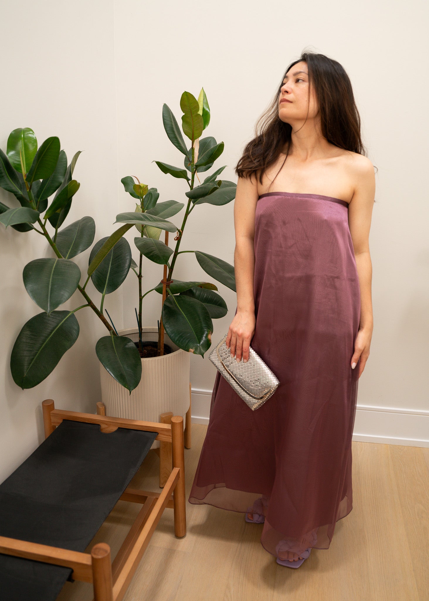 Woman in a strapless mauve purple dress holding a clutch, standing next to a plant indoors.