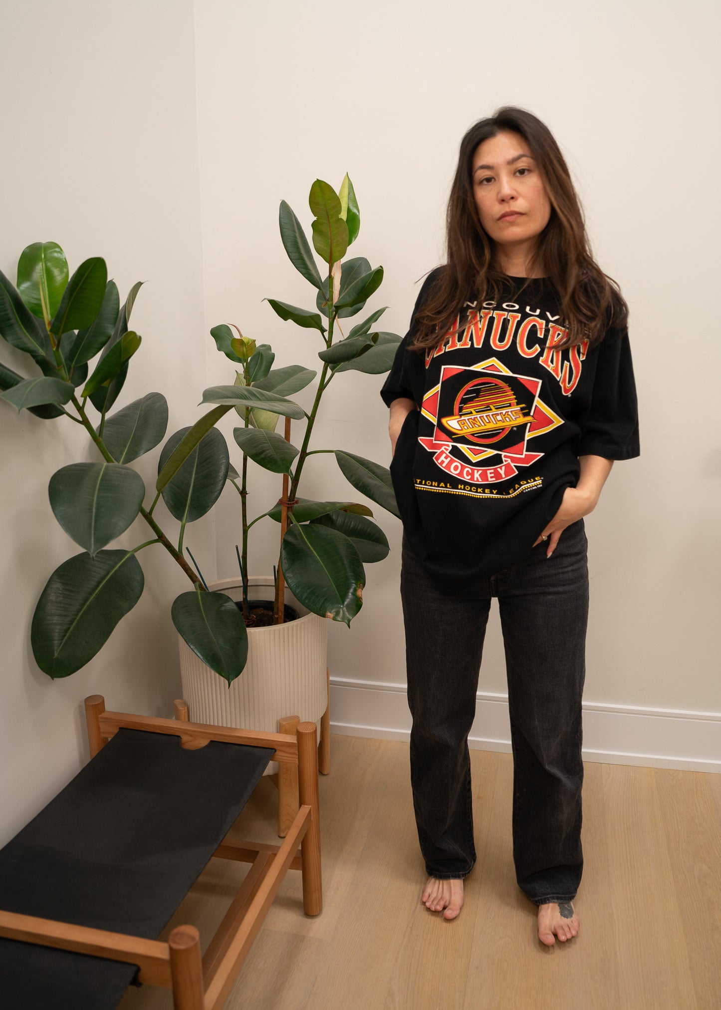 Person wearing a black t-shirt with a vintage Vancouver Canucks graphic design in a room with plants and a chair.