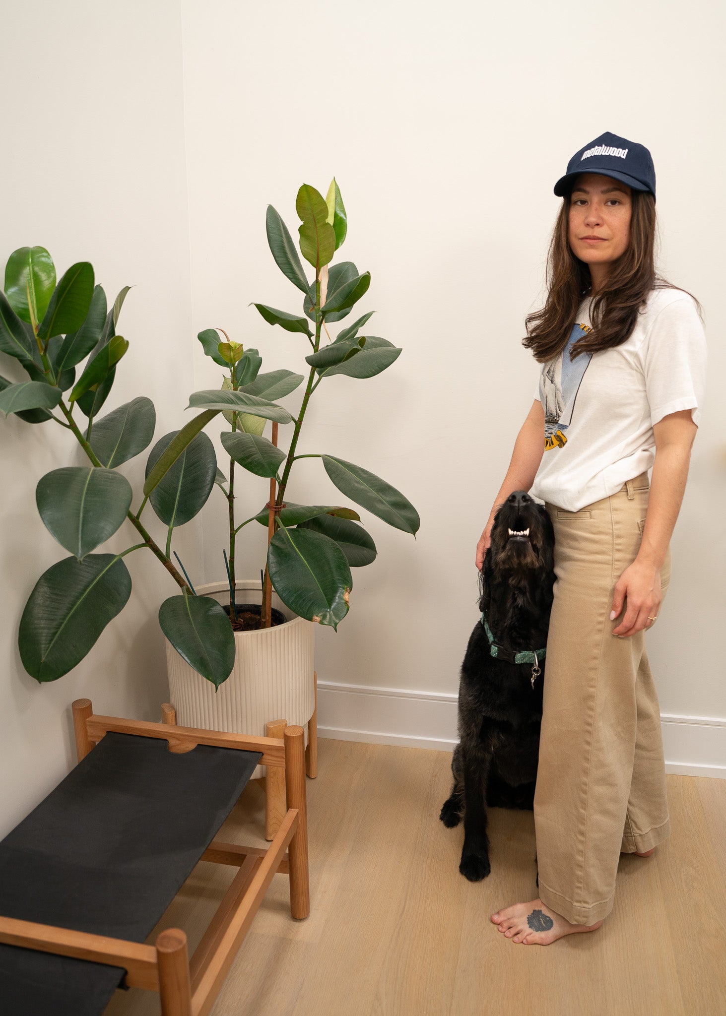 Woman wearing a boat graphic tee shirt standing next to a black dog in a room with plants and a small wooden table.