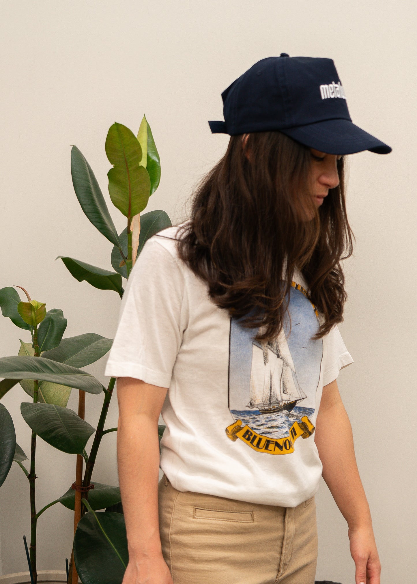 Person wearing a white t-shirt with a sailing graphic design and a navy cap, standing next to a plant.