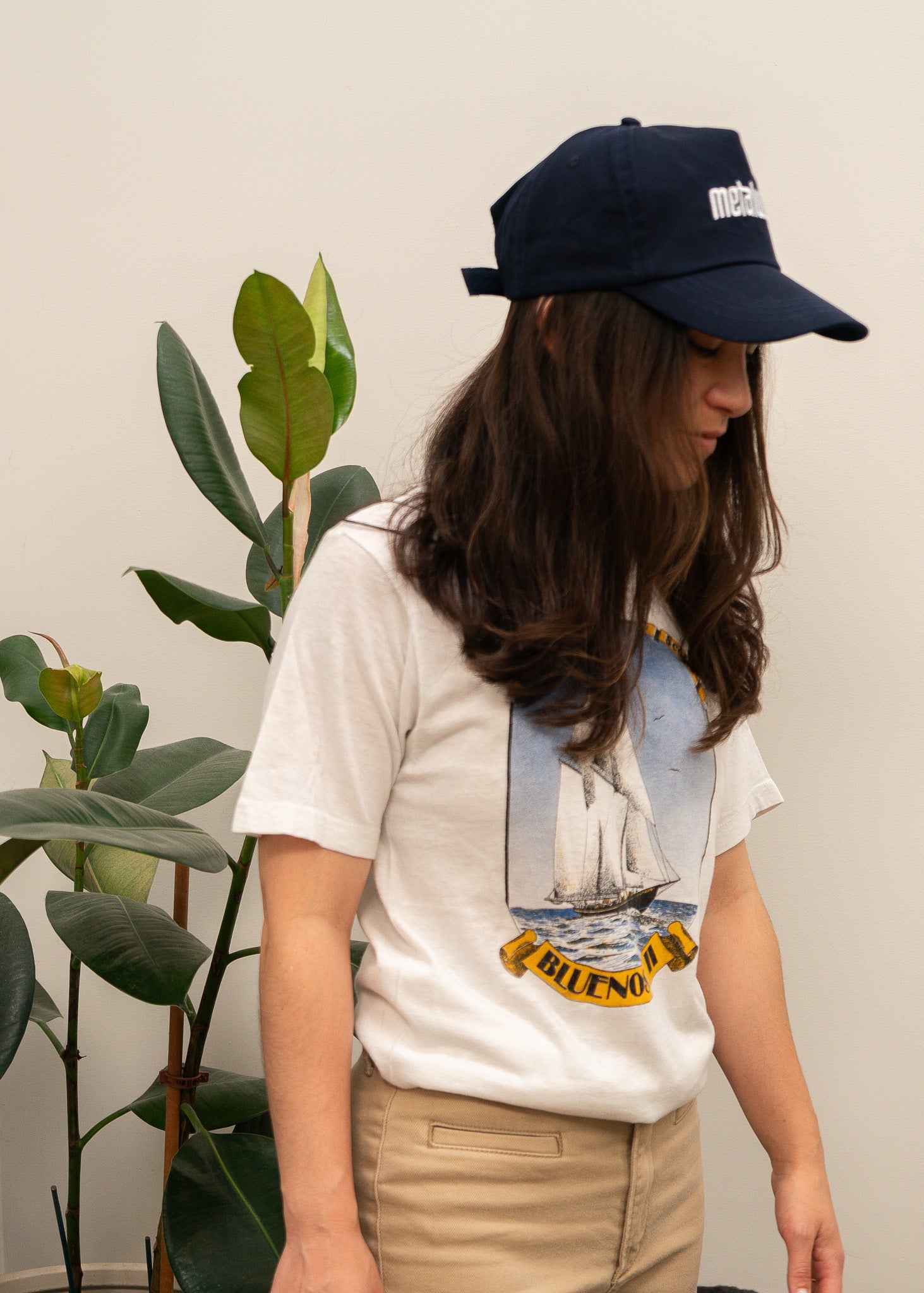 Person wearing a white t-shirt with a sailing graphic design and a navy cap, standing next to a plant.