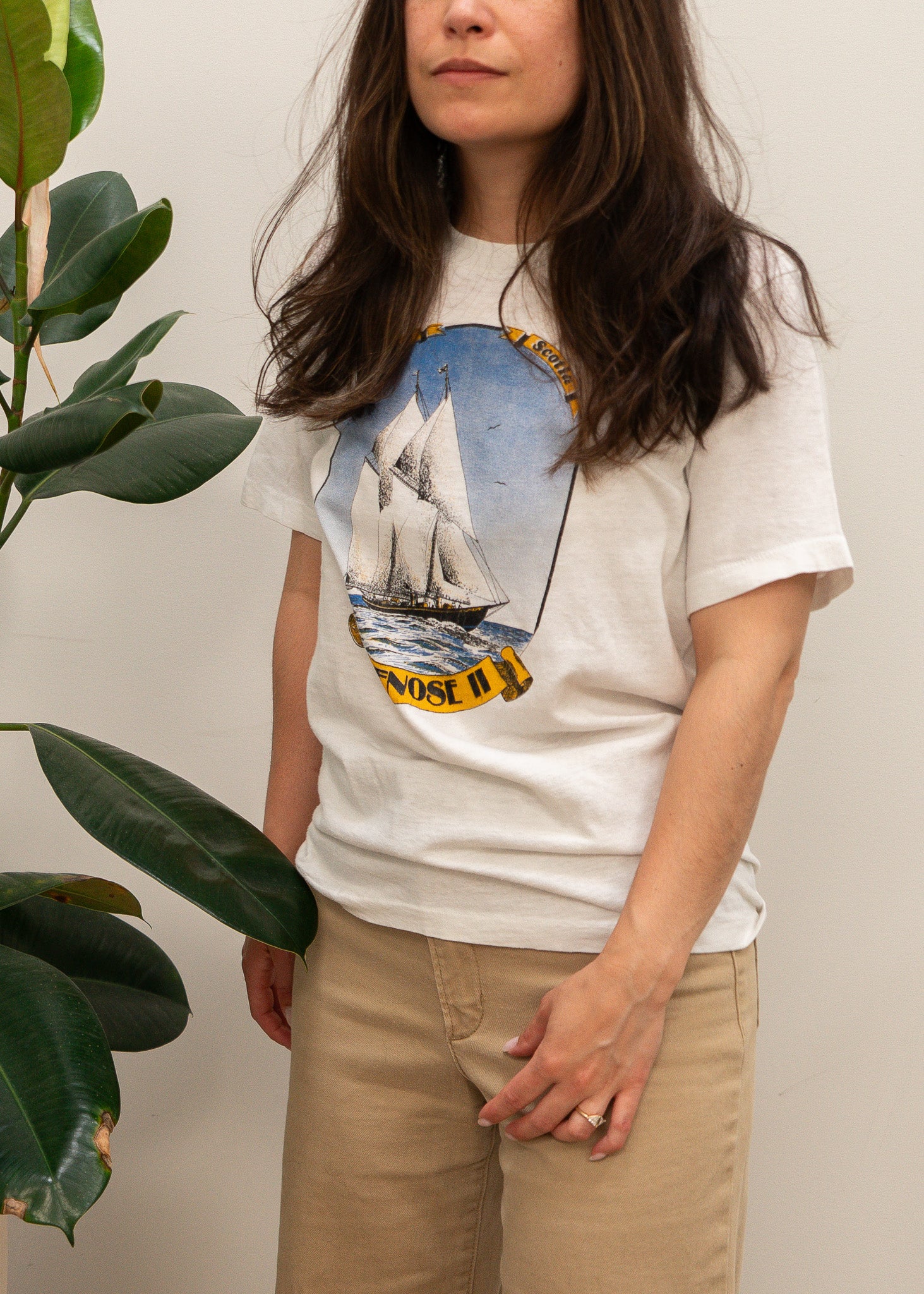 Person wearing a white t-shirt with a sailboat graphic, standing next to a plant.