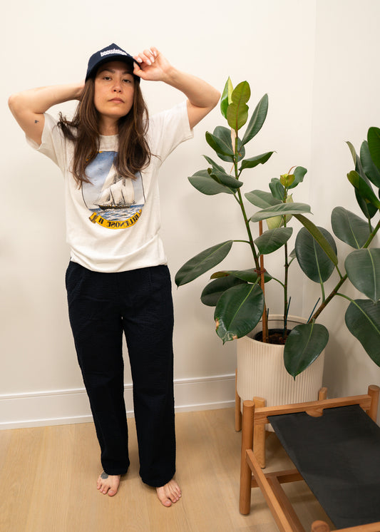 Person wearing a white t-shirt with a boat graphic design and dark pants, standing indoors with plants in the background.
