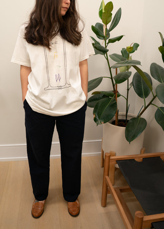 Person wearing a white t-shirt with a design and black pants standing indoors next to a plant.