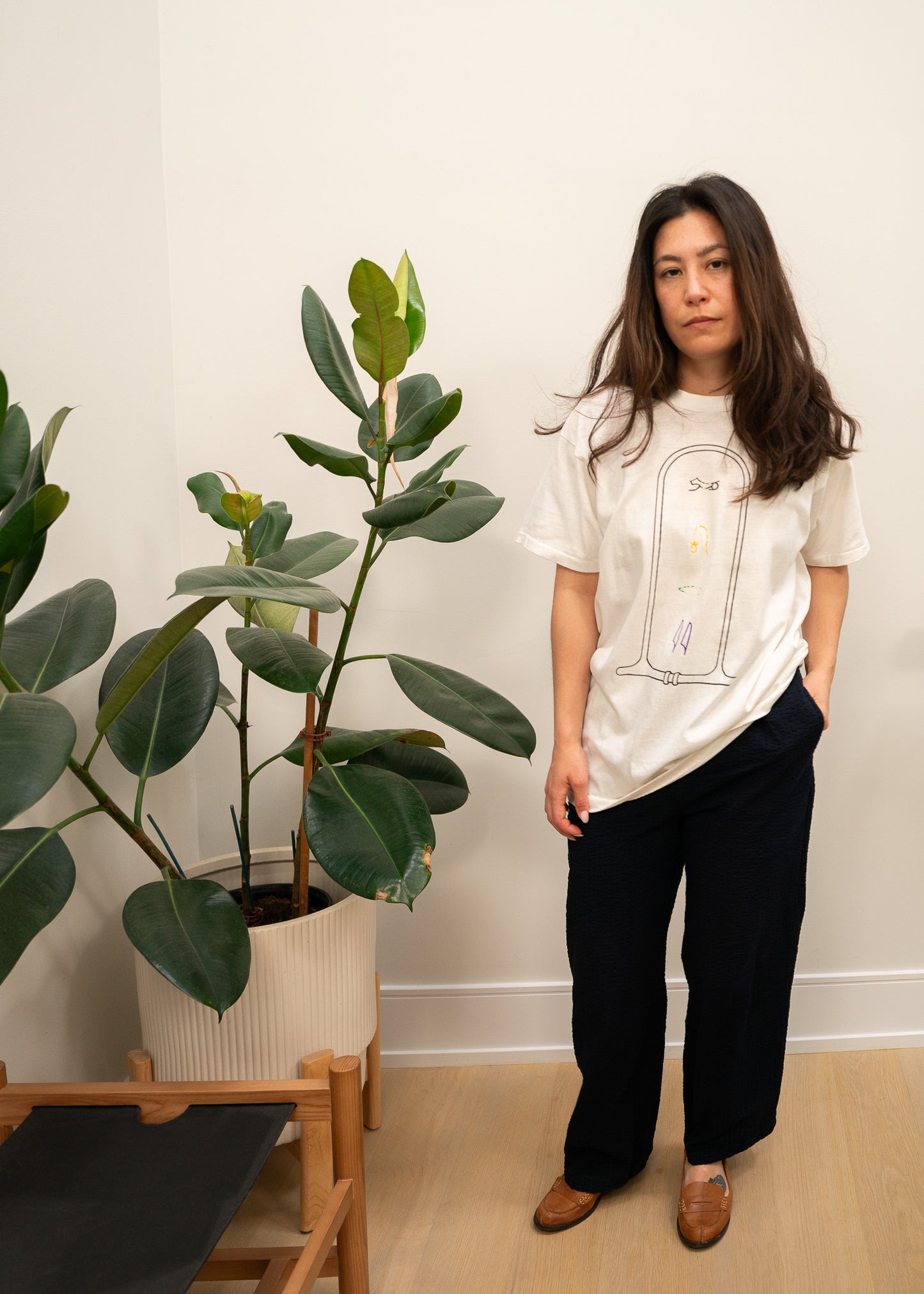 Person wearing a white t-shirt with a graphic design, standing next to a potted plant indoors.