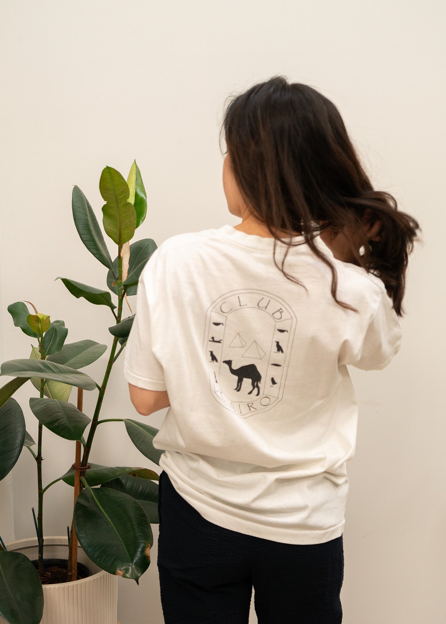 Person wearing a white t-shirt with Club Cairo a graphic design on the back, standing next to a potted plant.