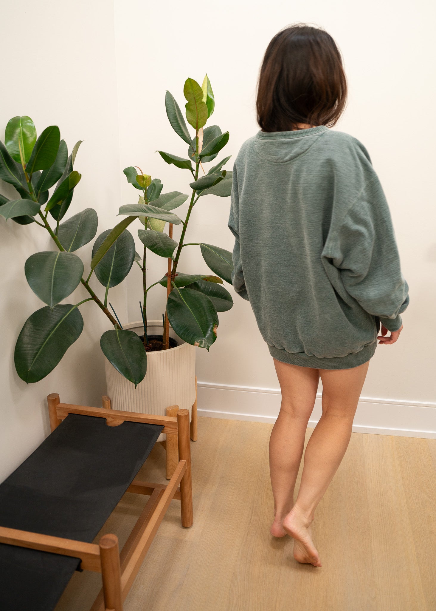 Person wearing a green oversized sweater standing in a room with plants and a wooden bench.