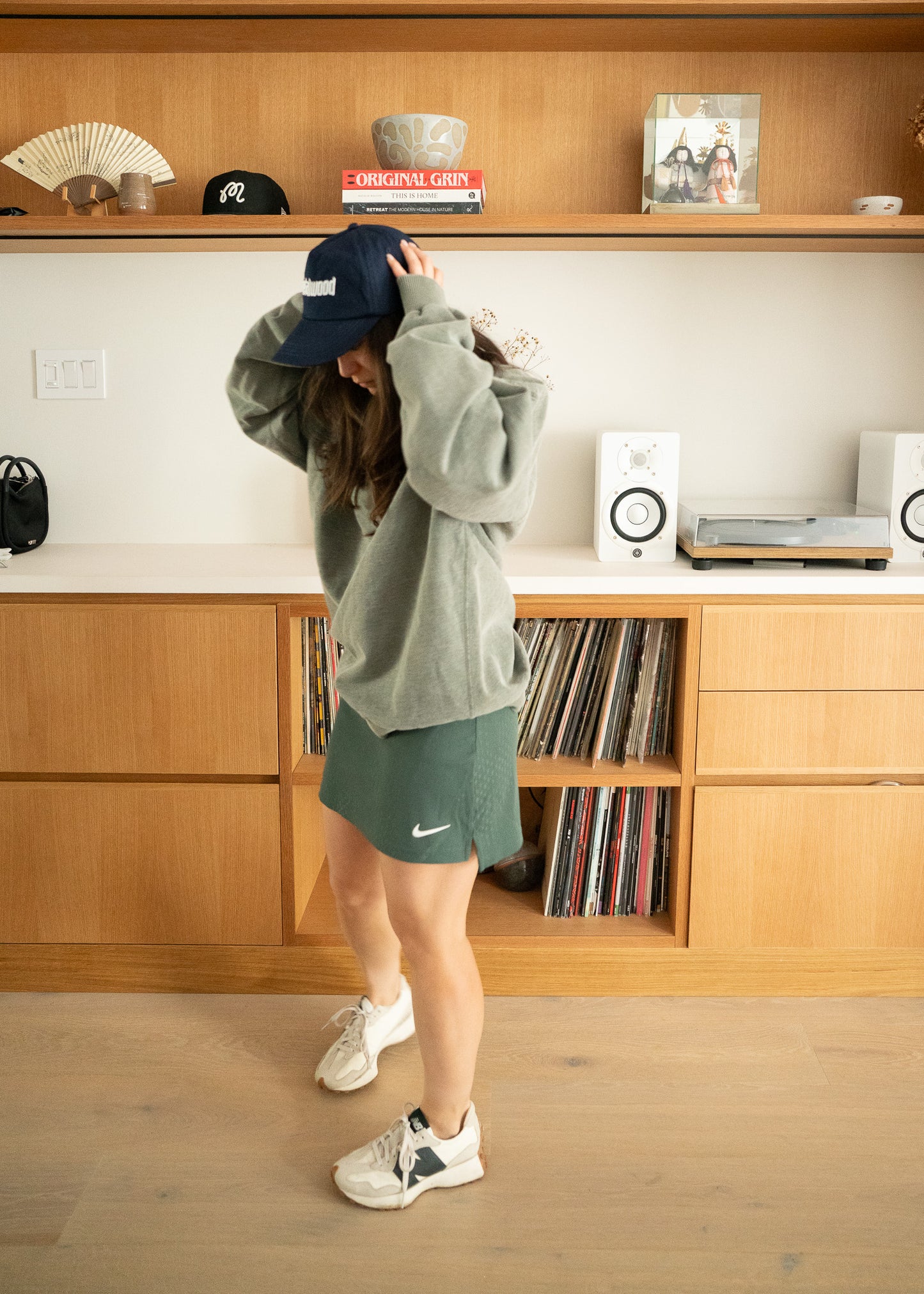 Person wearing an oversized green sweatshirt and Nike skirt in a room with wooden shelves and records.