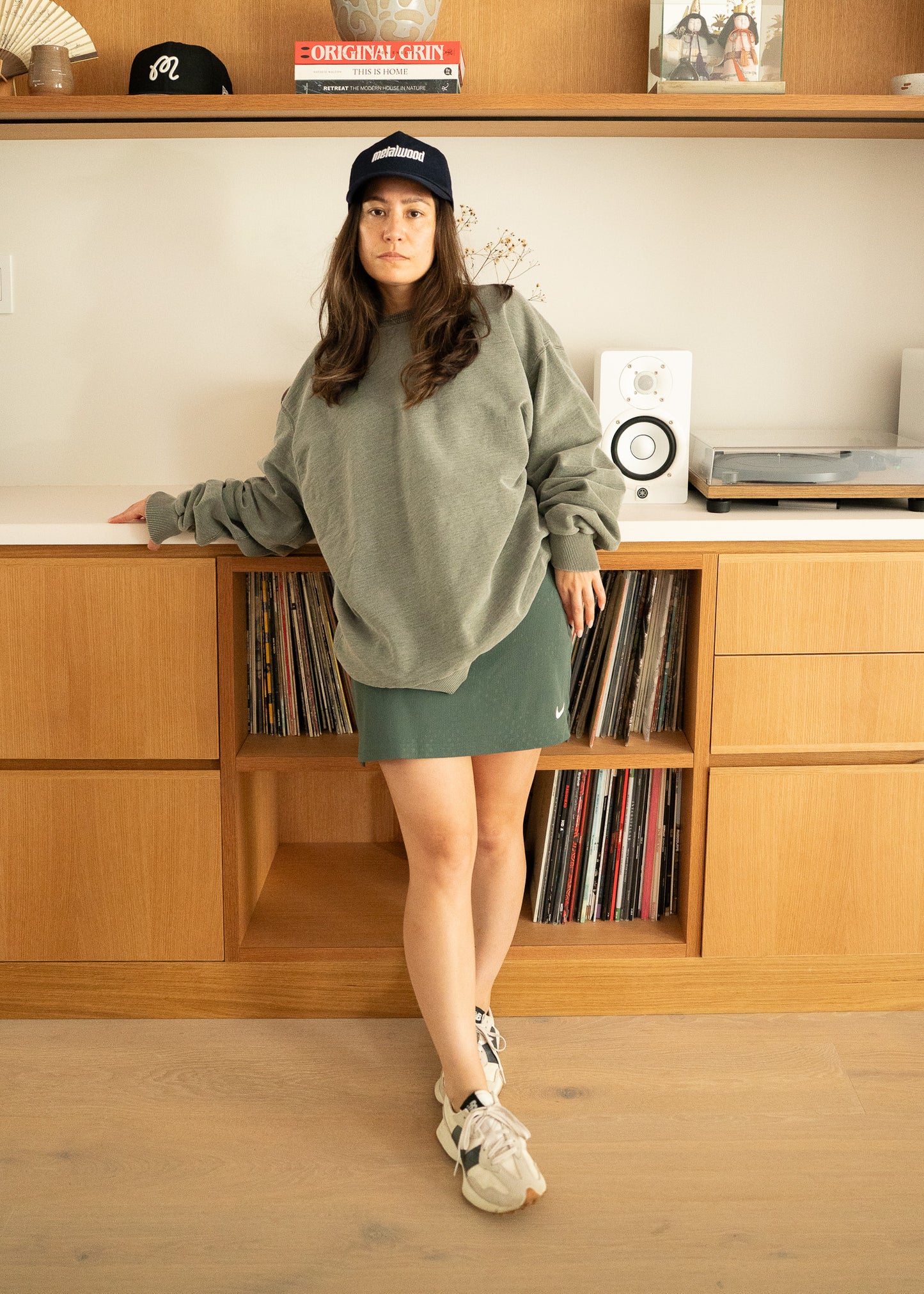 Woman in a green sweater and green skirt standing in front of a wooden shelf with records and speakers.