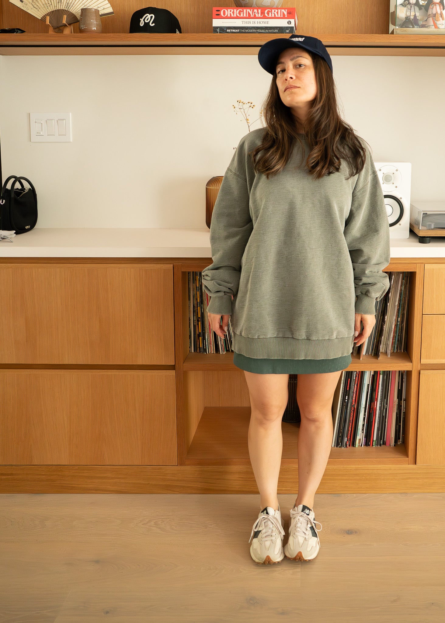 Person wearing an oversized green sweatshirt and dark cap indoors with wooden cabinets and books in the background